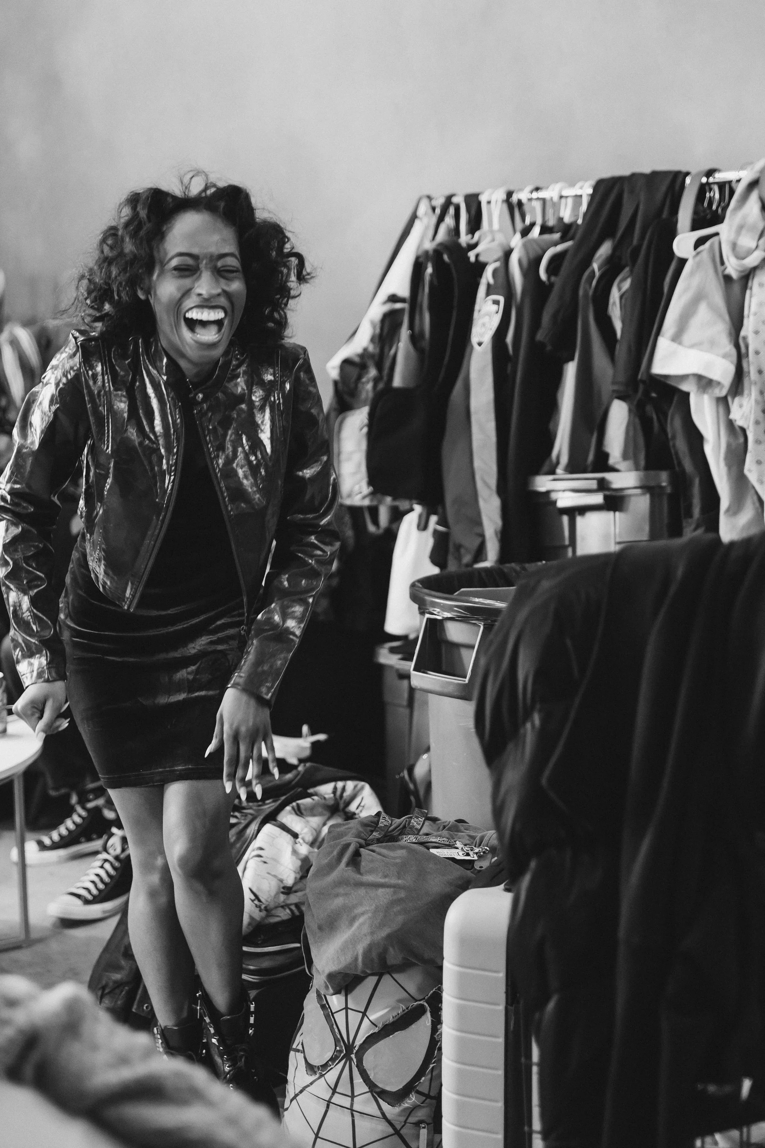 A woman with curly hair laughing or smiling, wearing a shiny jacket and a skirt, standing in what appears to be a cluttered laundry room or backstage area with clothes on racks and laundry baskets around.