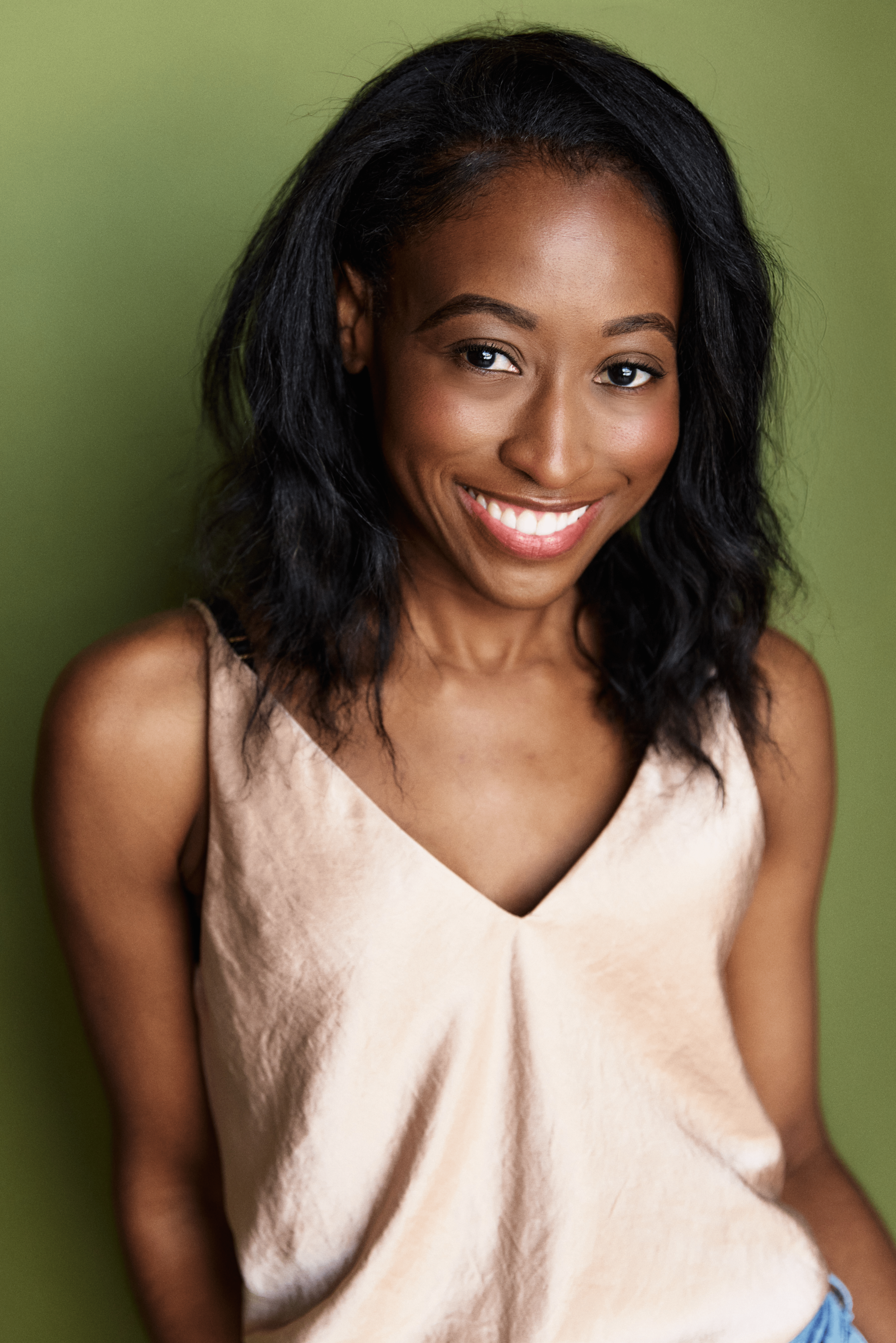 A young woman with black, wavy hair smiling and looking at the camera, wearing a beige sleeveless top, standing against a light green background.