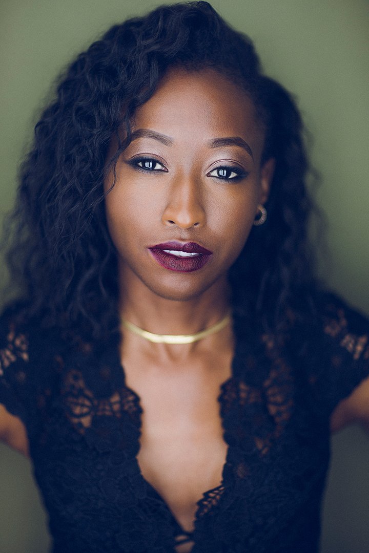 Portrait of a woman with curly hair and dark lipstick, wearing a lace top and gold choker necklace, against a green background.