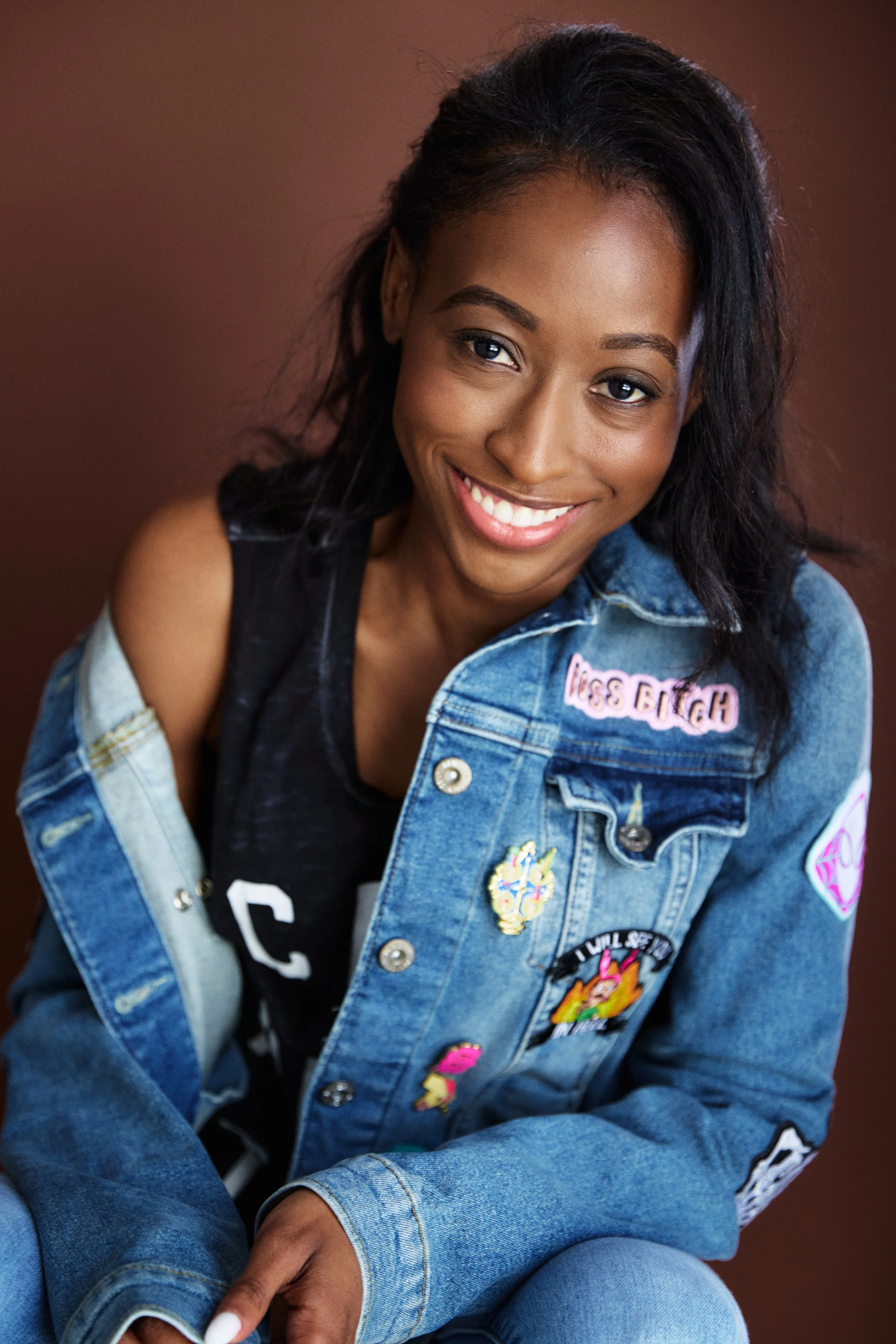 A young woman with dark curly hair and a radiant smile, wearing a black top and a denim jacket with patches and embroideries, sitting against a brown background.