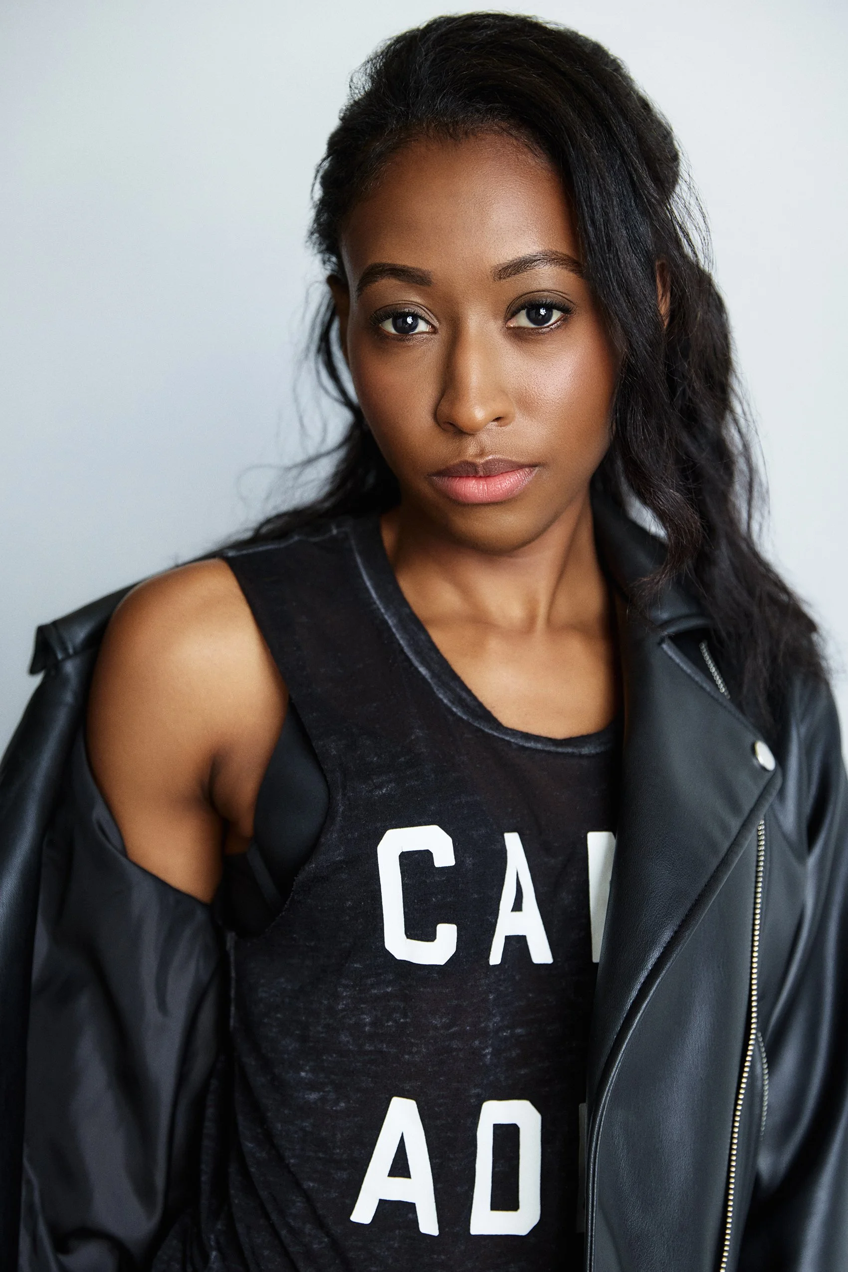 A young woman with dark hair and brown skin wearing a black tank top with white lettering and a black leather jacket draped over her shoulders.