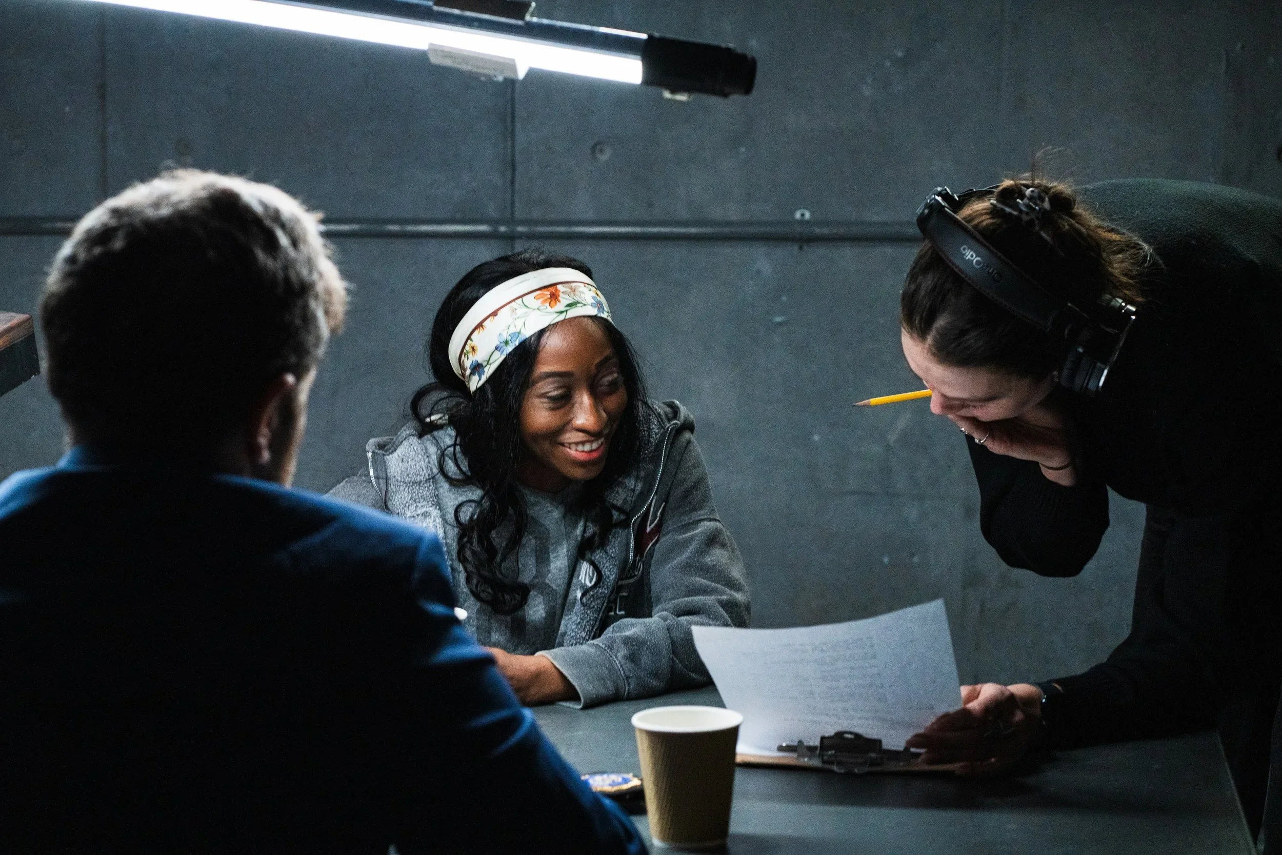 Three people sitting at a table, with one woman smiling, another person partially visible in the foreground, and a woman with headphones leaning over reading a document, in an indoor setting.