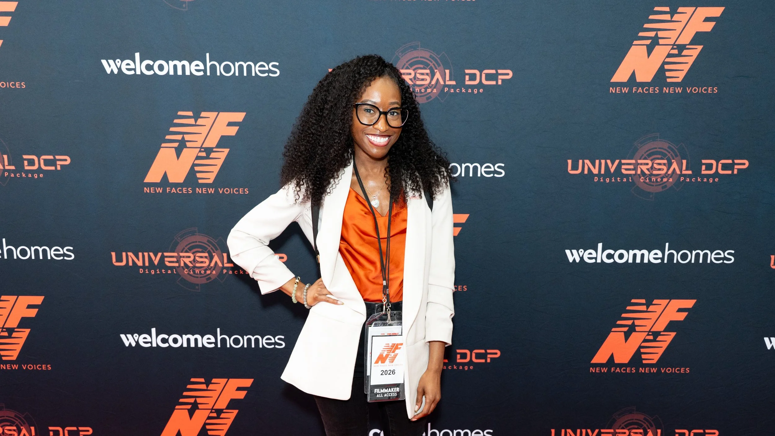 Asia Gordon smiles on the red carpet wearing a white blazer and filmmaker credentials at the New Faces New Voices Film Festival.