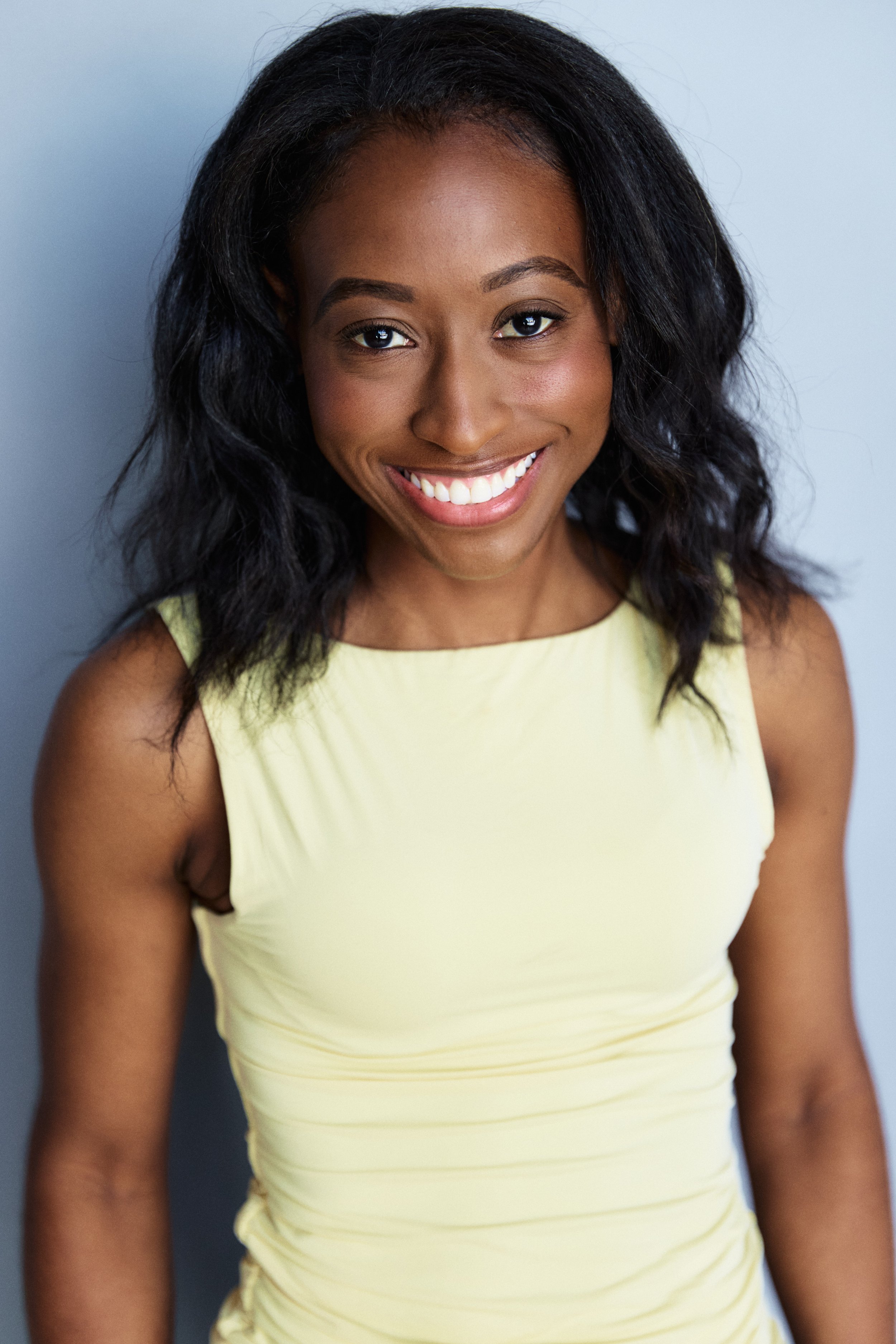A young woman with dark, curly hair smiling, wearing a sleeveless yellow top, standing against a light gray background.
