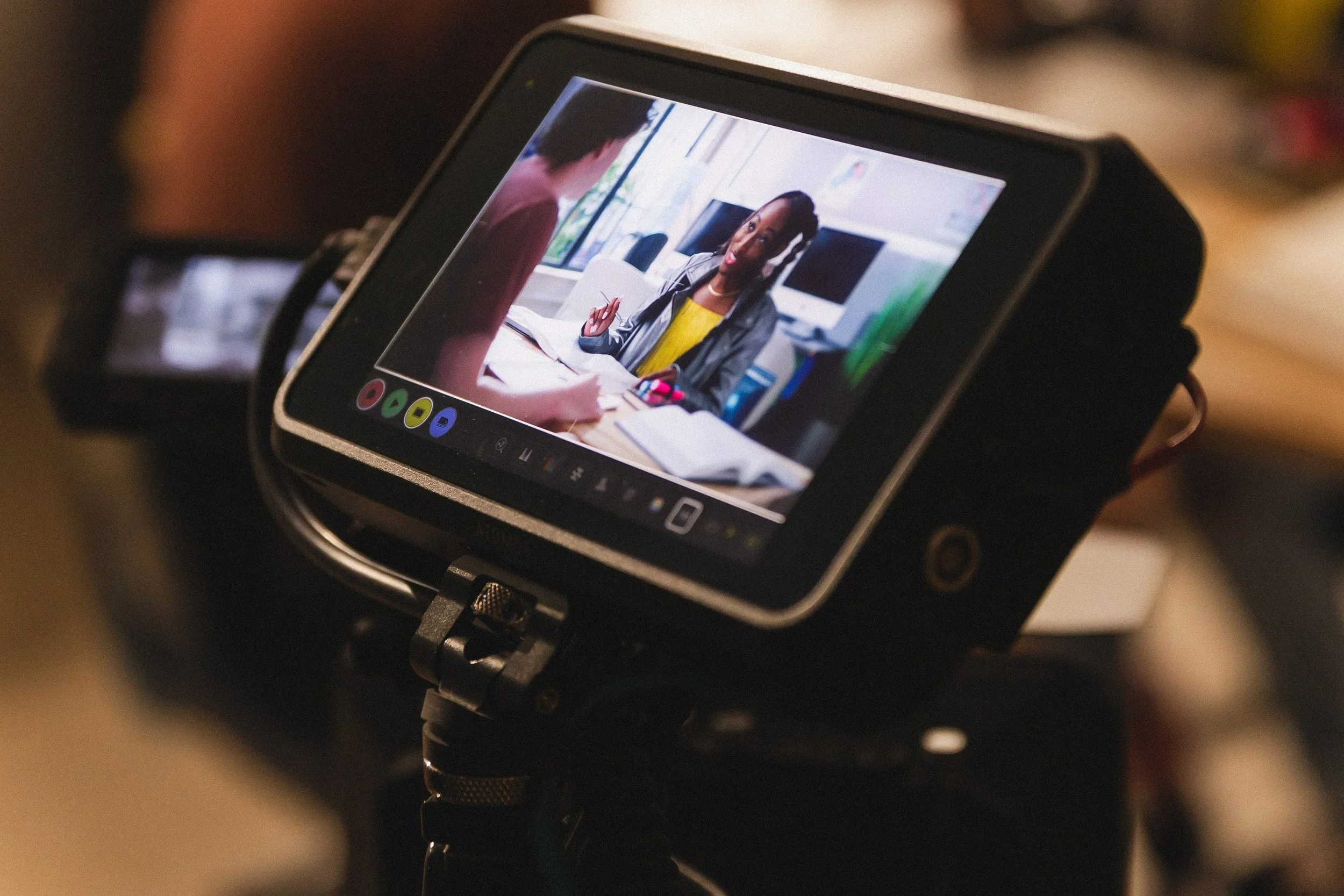 A camera viewfinder showing a woman with dark skin and braids, sitting at a desk with a notebook and pen, engaged in a conversation with a person whose back is to the camera in an office with large windows and monitors.