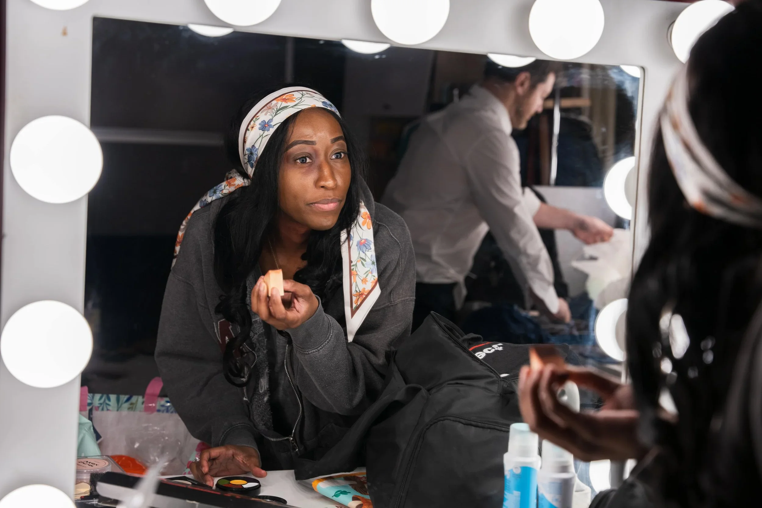 A woman with a floral headscarf and a gray hoodie sitting at a dressing room mirror, applying makeup with various makeup items on the table in front of her.