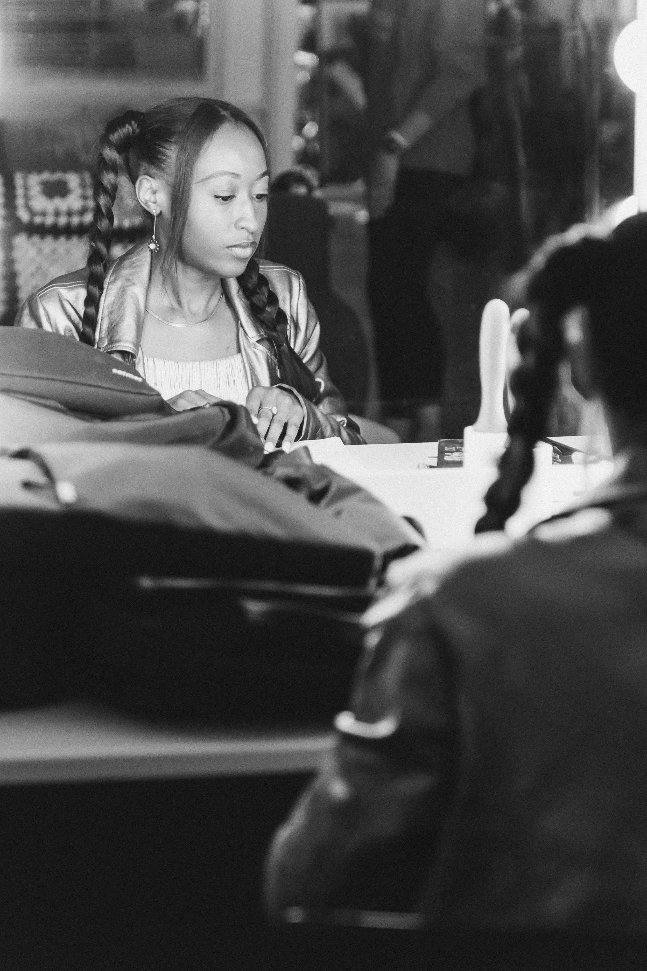 A young woman with braided hair and earrings, sitting at a table in a restaurant or cafe, looking down, with a blurred background of other people and the interior.