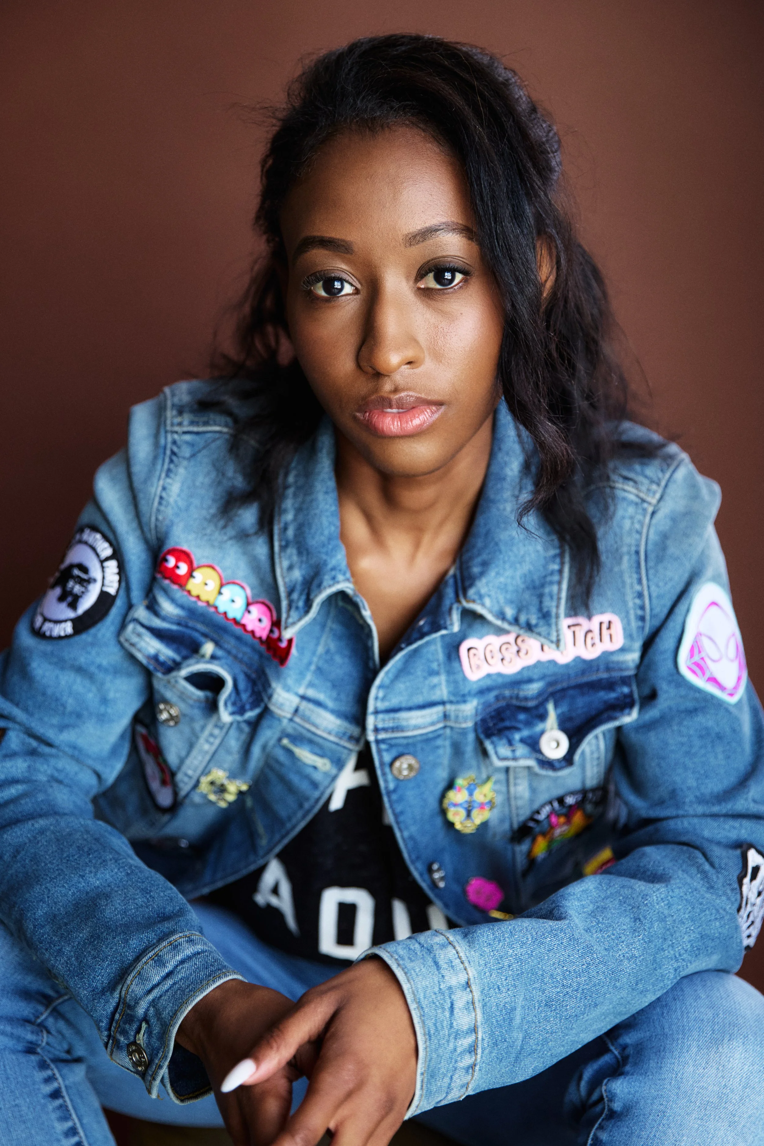 A young woman with dark curly hair and medium brown skin, looking directly into the camera, wearing a blue denim jacket decorated with various patches and pins, sitting against a brown background.