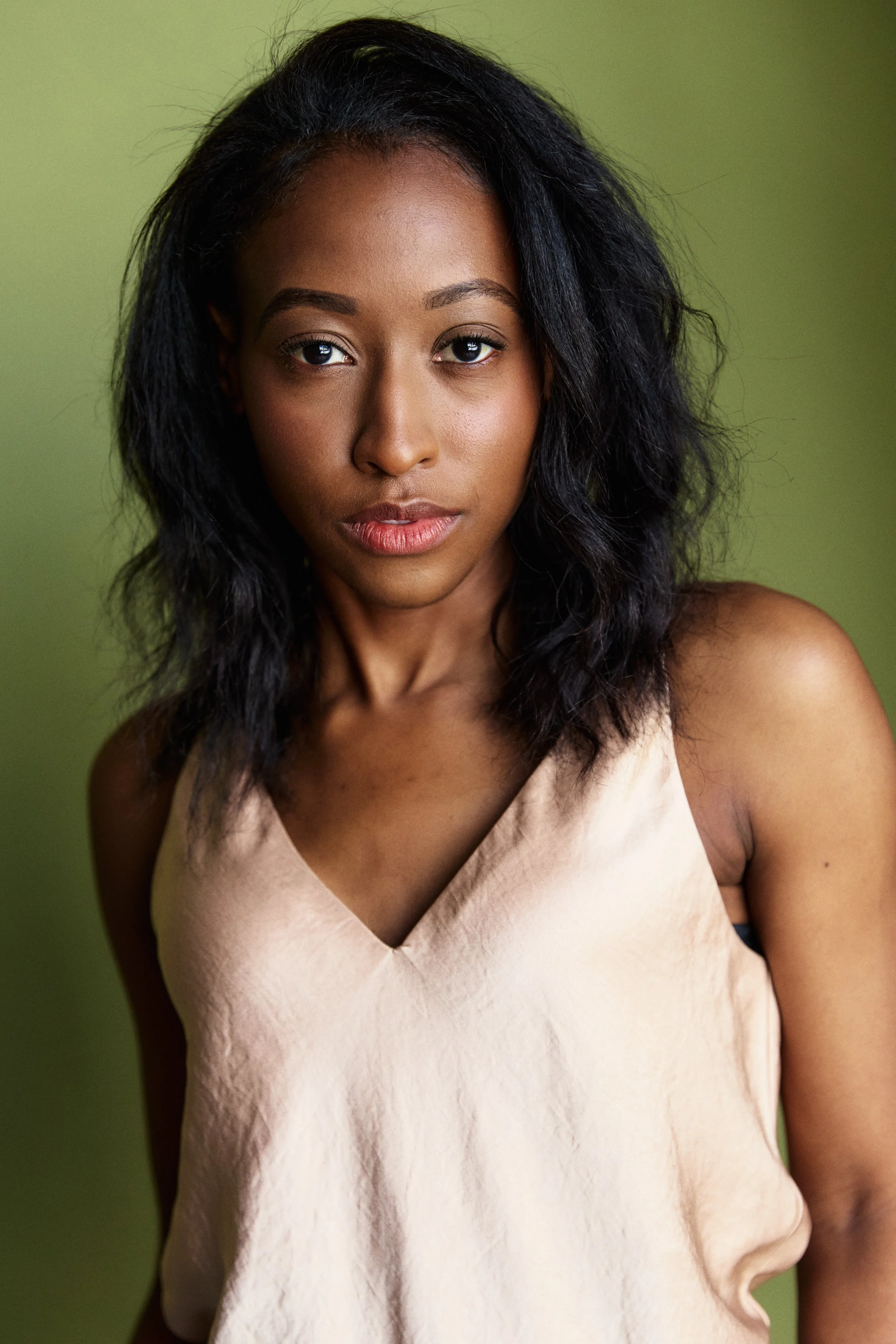A young woman with dark, wavy hair and medium skin, looking directly at the camera with a neutral expression, wearing a sleeveless beige top, against a light green background.