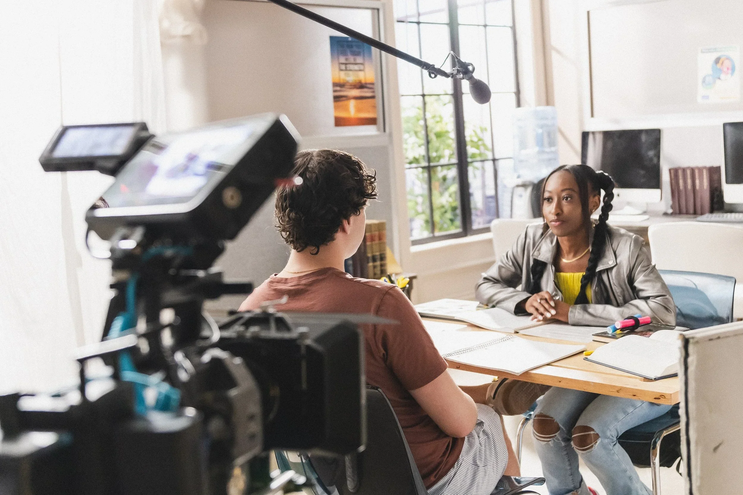 A woman with braids and a yellow shirt talks to a person in a brown t-shirt during an interview or conversation, with filming equipment visible in the foreground, in a bright room with large windows.