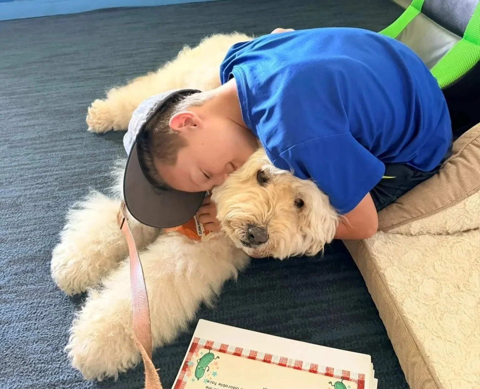 A young boy in a blue shirt and gray hat cuddling a large cream-colored fluffy dog lying on the floor. The boy is laying on his side with his head pressed against the dog's head, and they are both resting their faces gently together. There is a piece of paper with a cartoon pickle on it in the foreground.