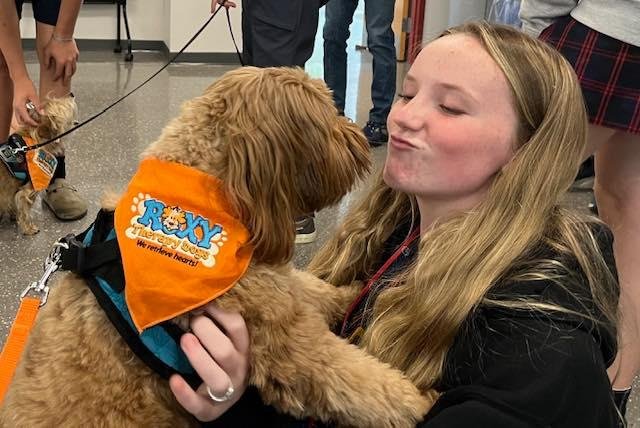 Girl with long blonde hair hugging a golden doodle wearing an orange bandana and a harness.