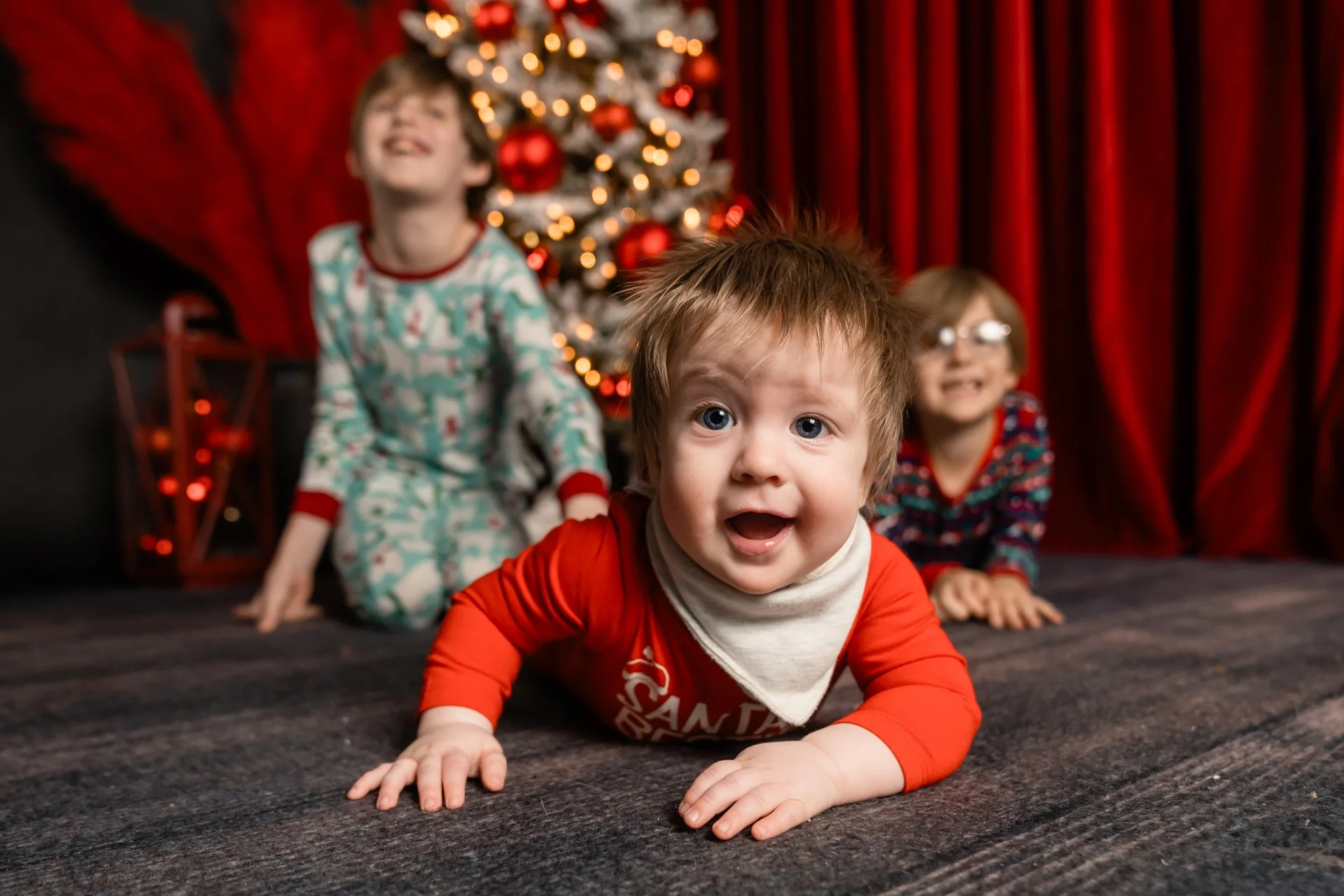 Three children in Christmas pajamas playing on the floor in front of a decorated Christmas tree with red ornaments, with red curtains in the background.