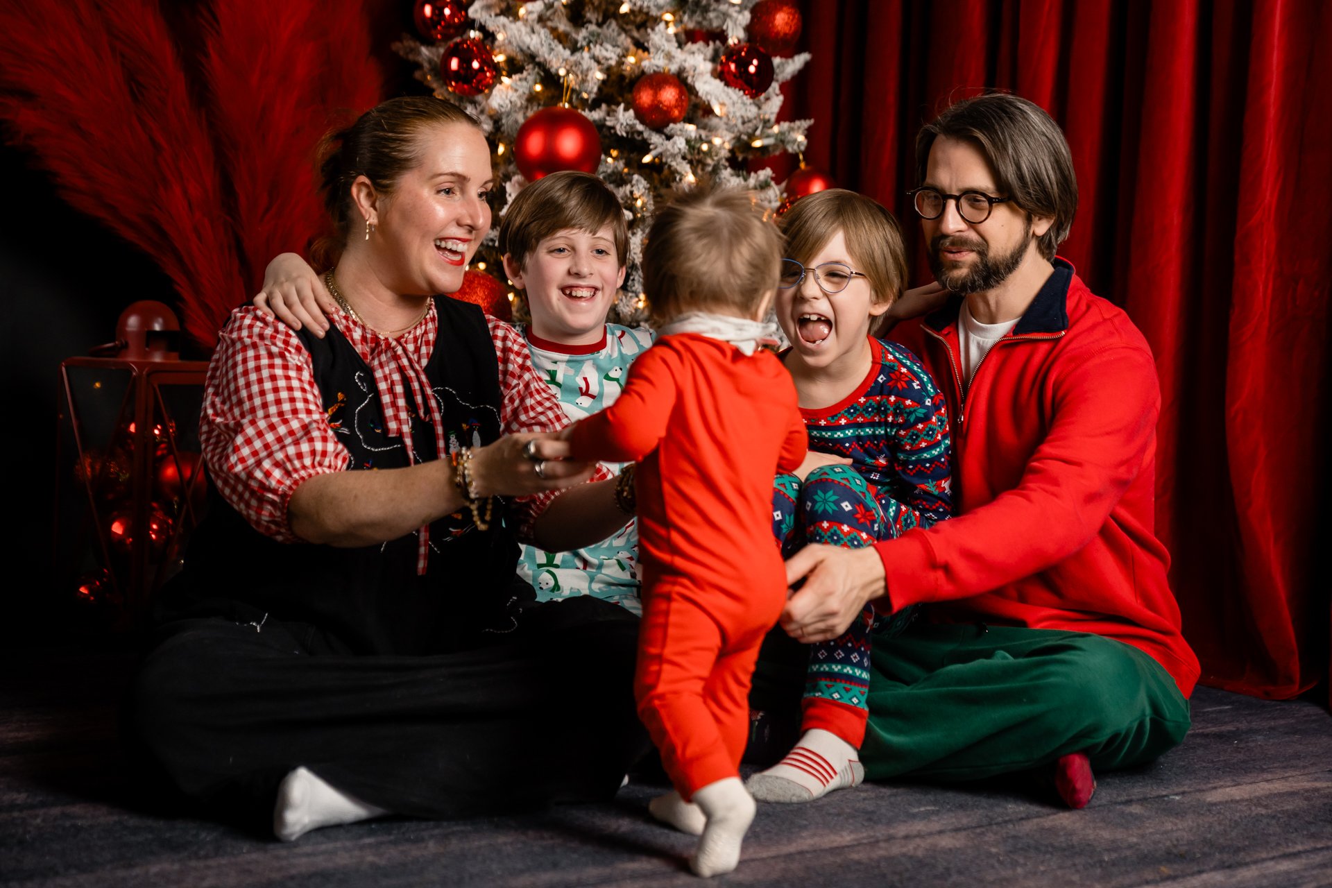 A family of six celebrating Christmas around a decorated Christmas tree, sitting on the floor in front of red curtains. The children and adults are smiling and laughing, with the youngest child in red standing and the others sitting, wearing festive pajamas.