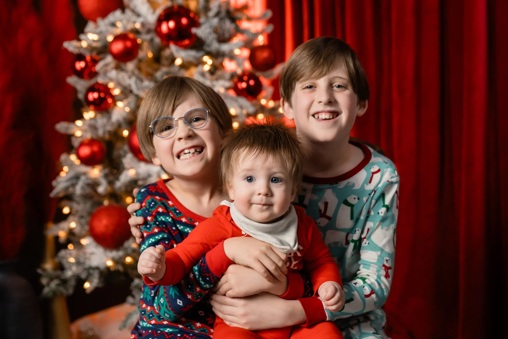 Three children smiling in front of a decorated Christmas tree with red ornaments, wearing Christmas pajamas.
