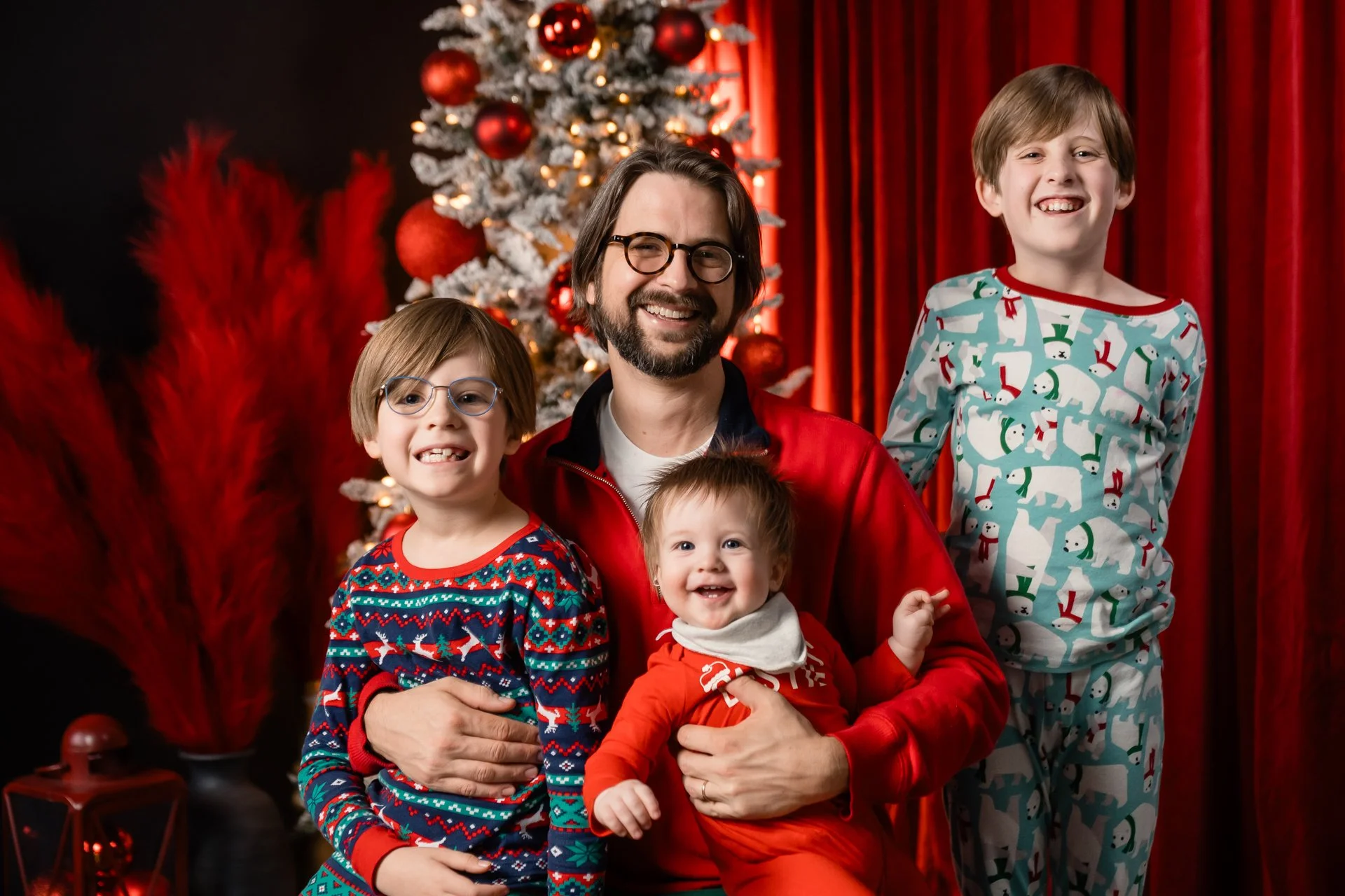 A man with glasses and a beard wearing a red jacket, sitting with three children in pajamas in front of a Christmas tree and red curtains. The children are smiling, with one in a Christmas sweater, one in Christmas pajamas, and an infant in red outfit.