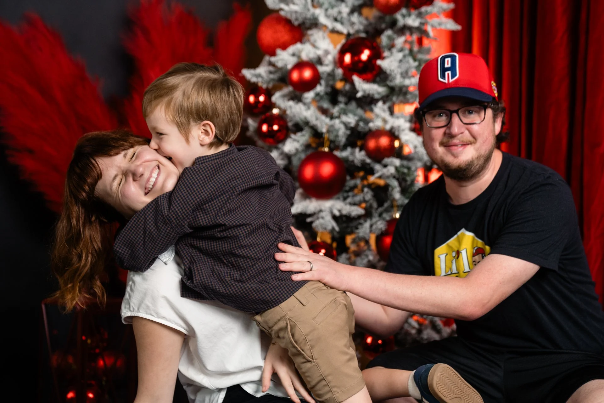 A family of three celebrating Christmas in front of a decorated tree, with a woman and a young boy hugging and a man smiling at the camera.