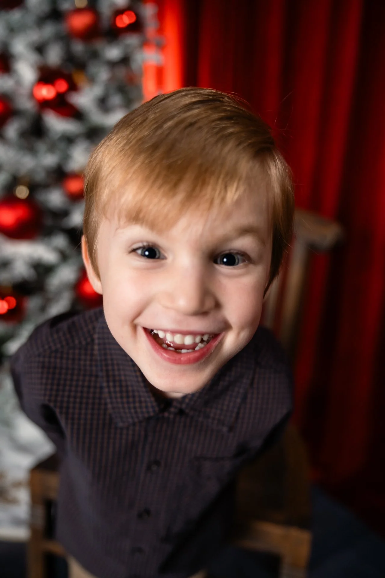 Close-up of a happy young boy with red hair and blue eyes, smiling broadly, standing in front of a decorated Christmas tree with red ornaments.