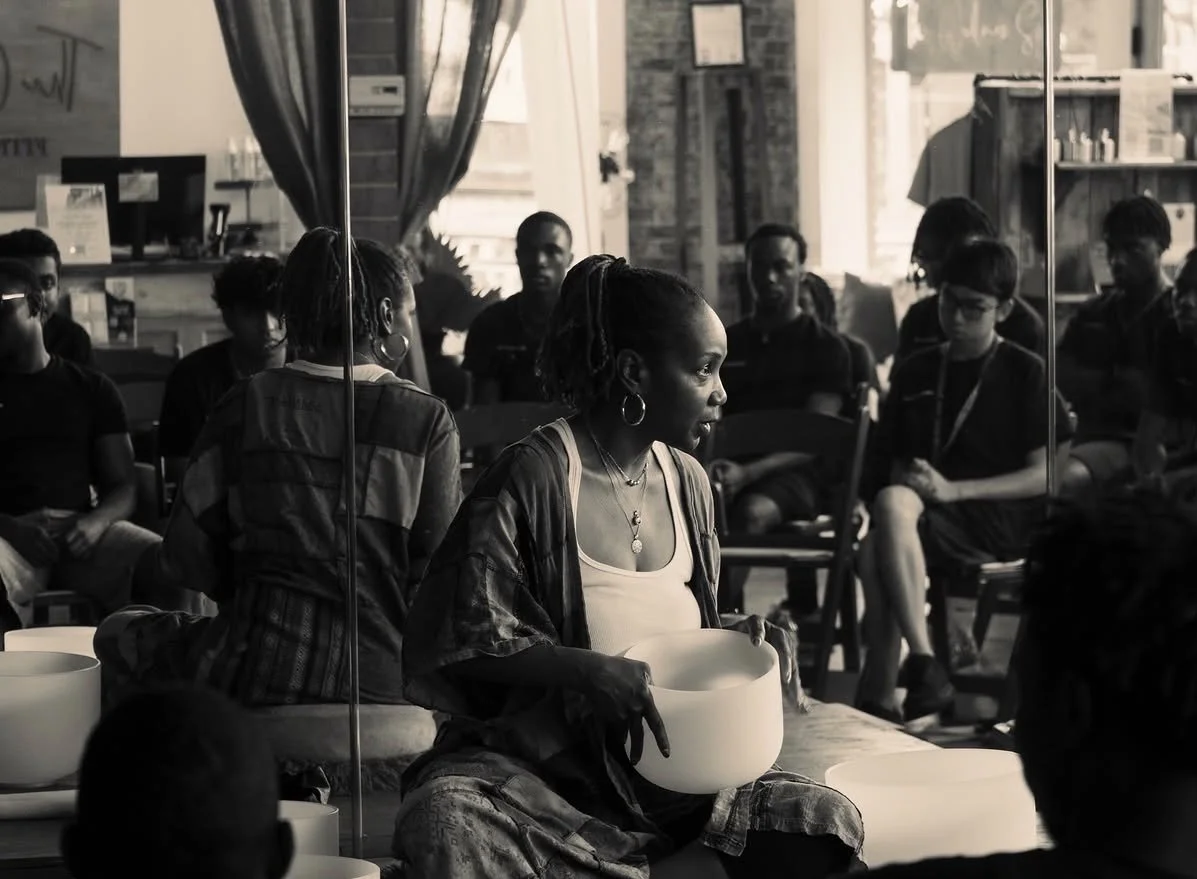 A woman holding a singing bowl surrounded by people sitting in a semi-circle in a cozy indoor space with large windows and brick walls.