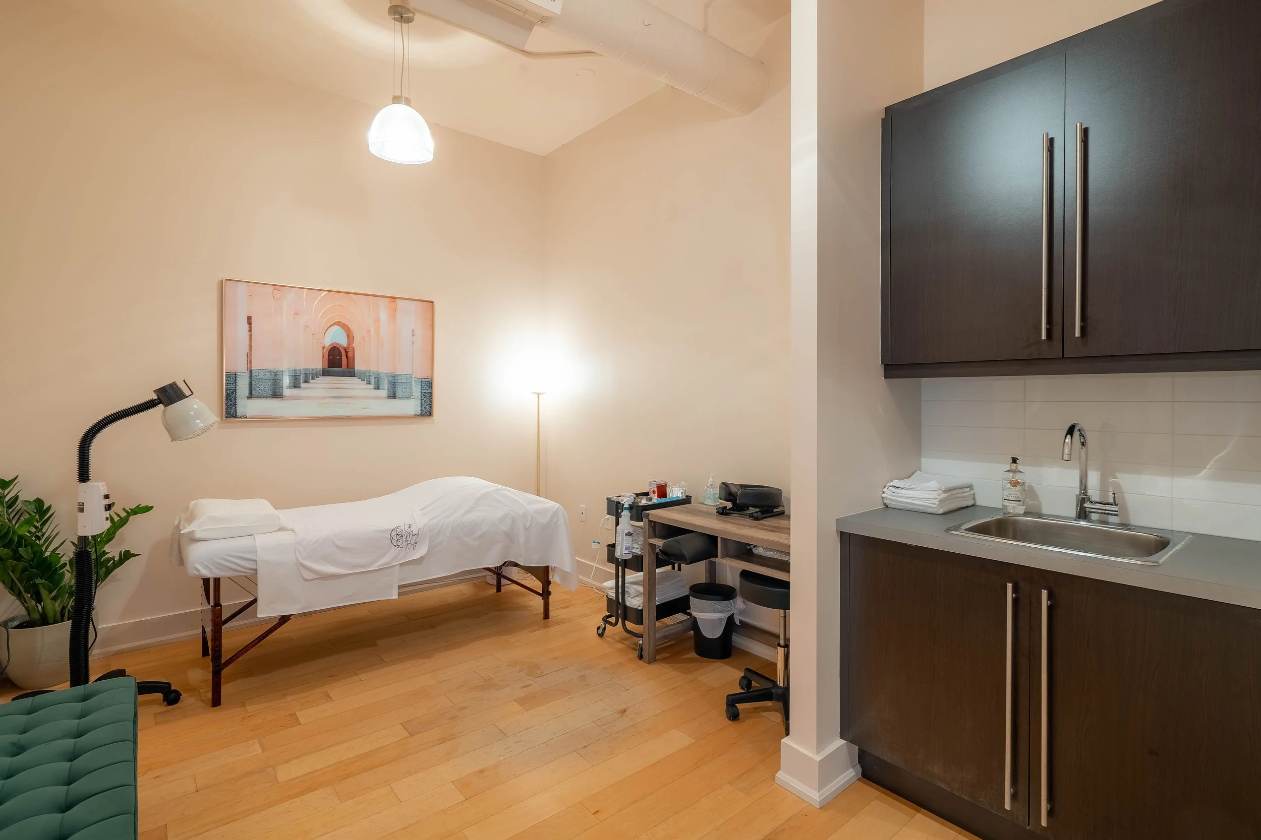 A massage room with a massage table covered in white sheet, a wall painting of arches, a standing lamp, a cart with massage supplies, a bathroom sink with soap, stacked towels, dark wooden cabinets, a green bench, and wooden flooring.