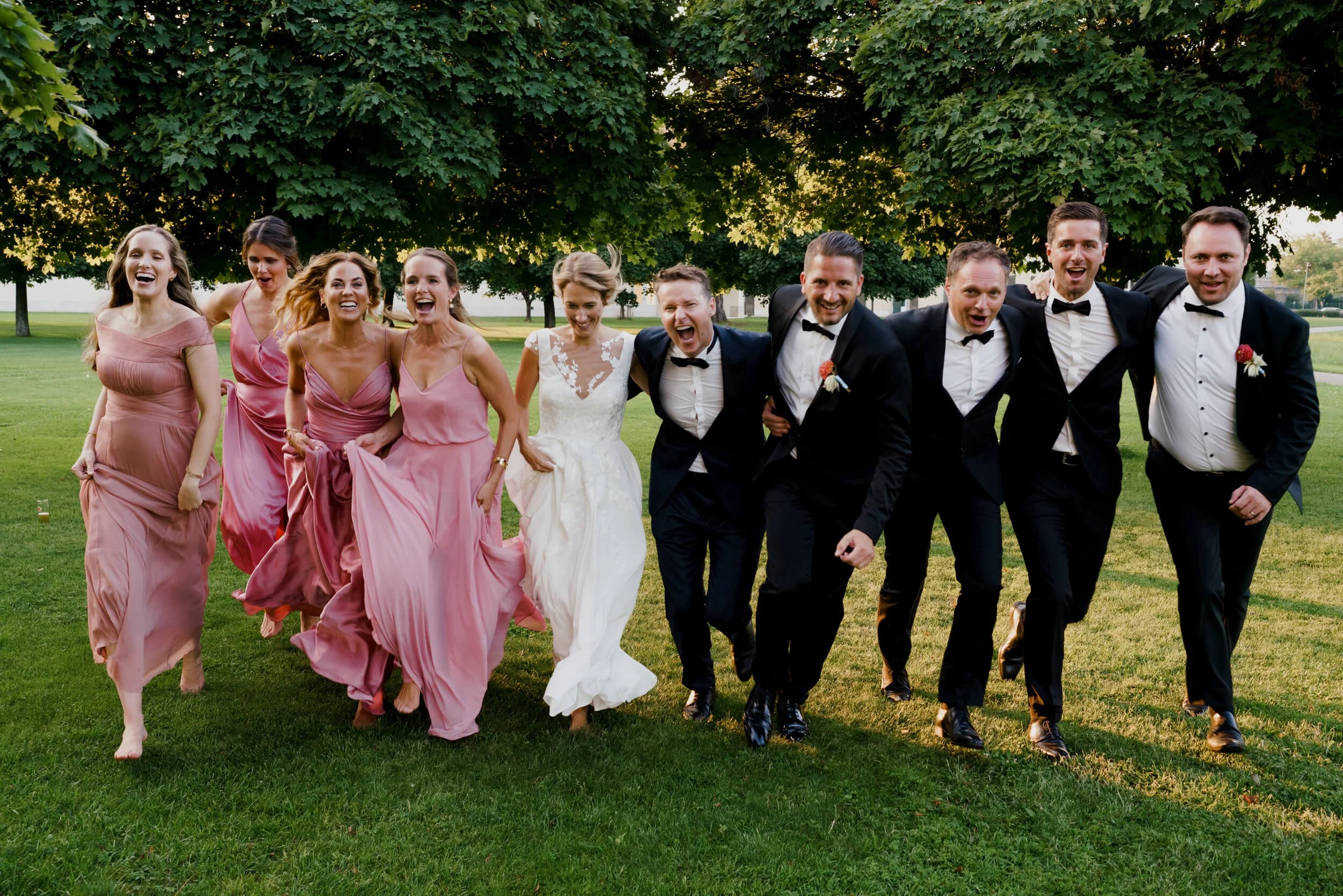 Wedding couple is running toward camera of wedding photographer together with bridesmaids in black tie outfits and pink dresses
