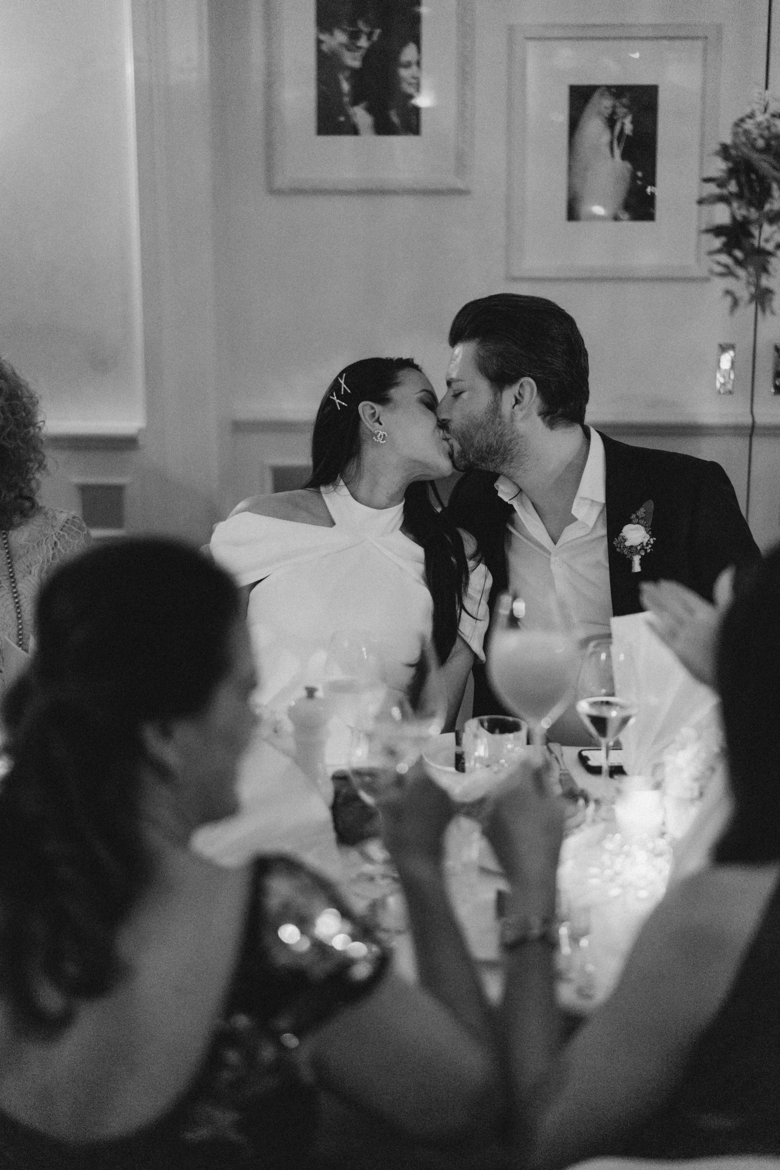 A couple kissing at a wedding reception, surrounded by seated guests and drinks on the table.