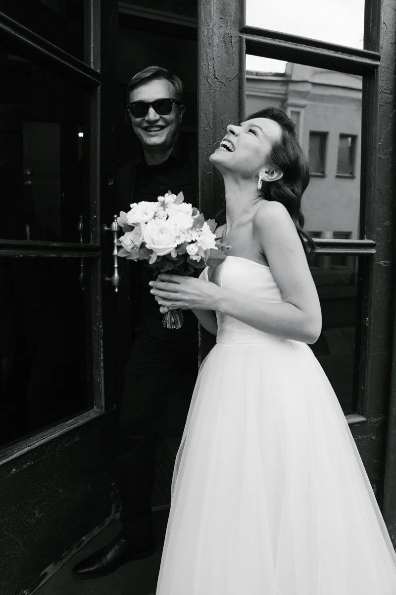 Black and white photo of modern wedding couple, bride is laughing holding her bouquet, groom is standing in the doorframe in sunglasses