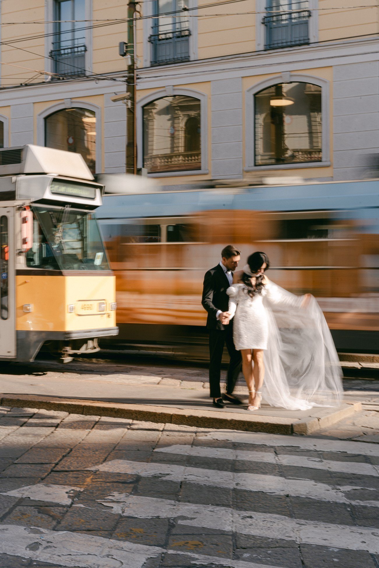 A couple dressed in wedding attire standing on a city street corner with a tram passing by behind them.
