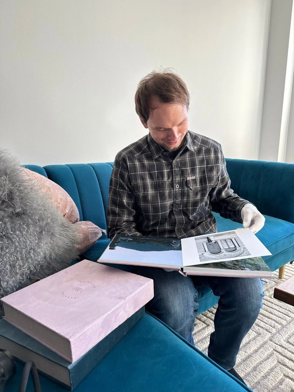 A young man with brown hair wearing a black and gray plaid shirt sits on a blue velvet couch, looking at a photo book with pictures. There is a pink box on the couch next to him.