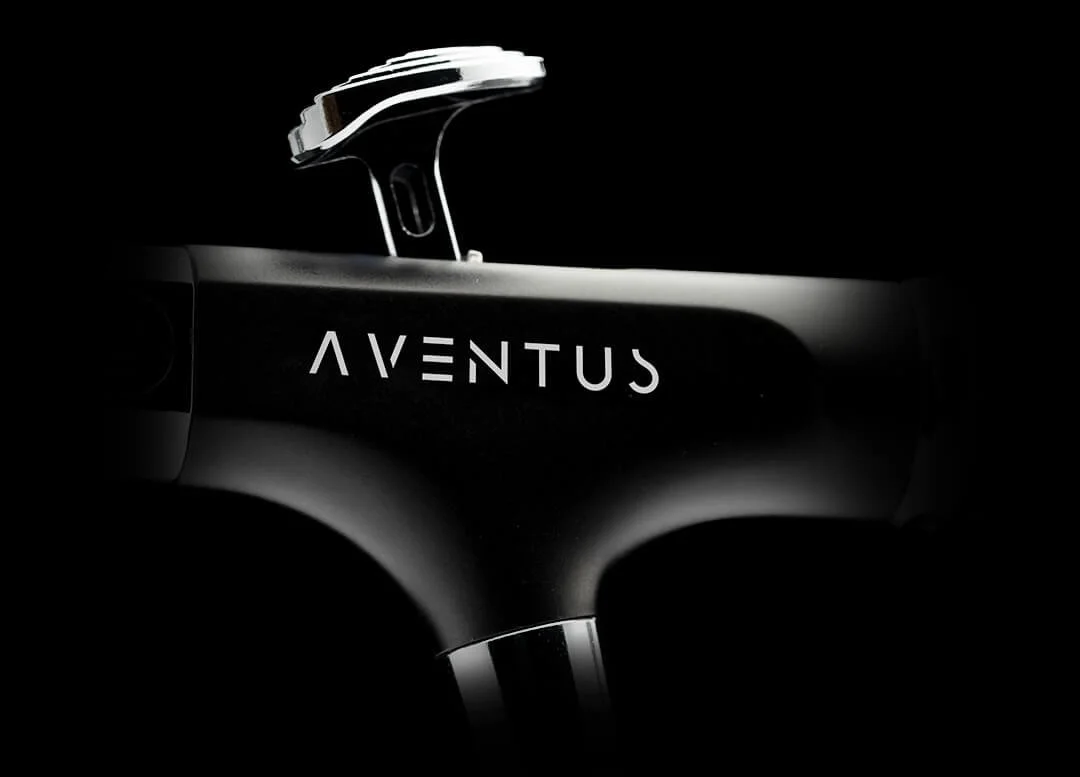 Close-up of a black electric skateboard, with the brand name 'Aventus' written on it, against a dark background.