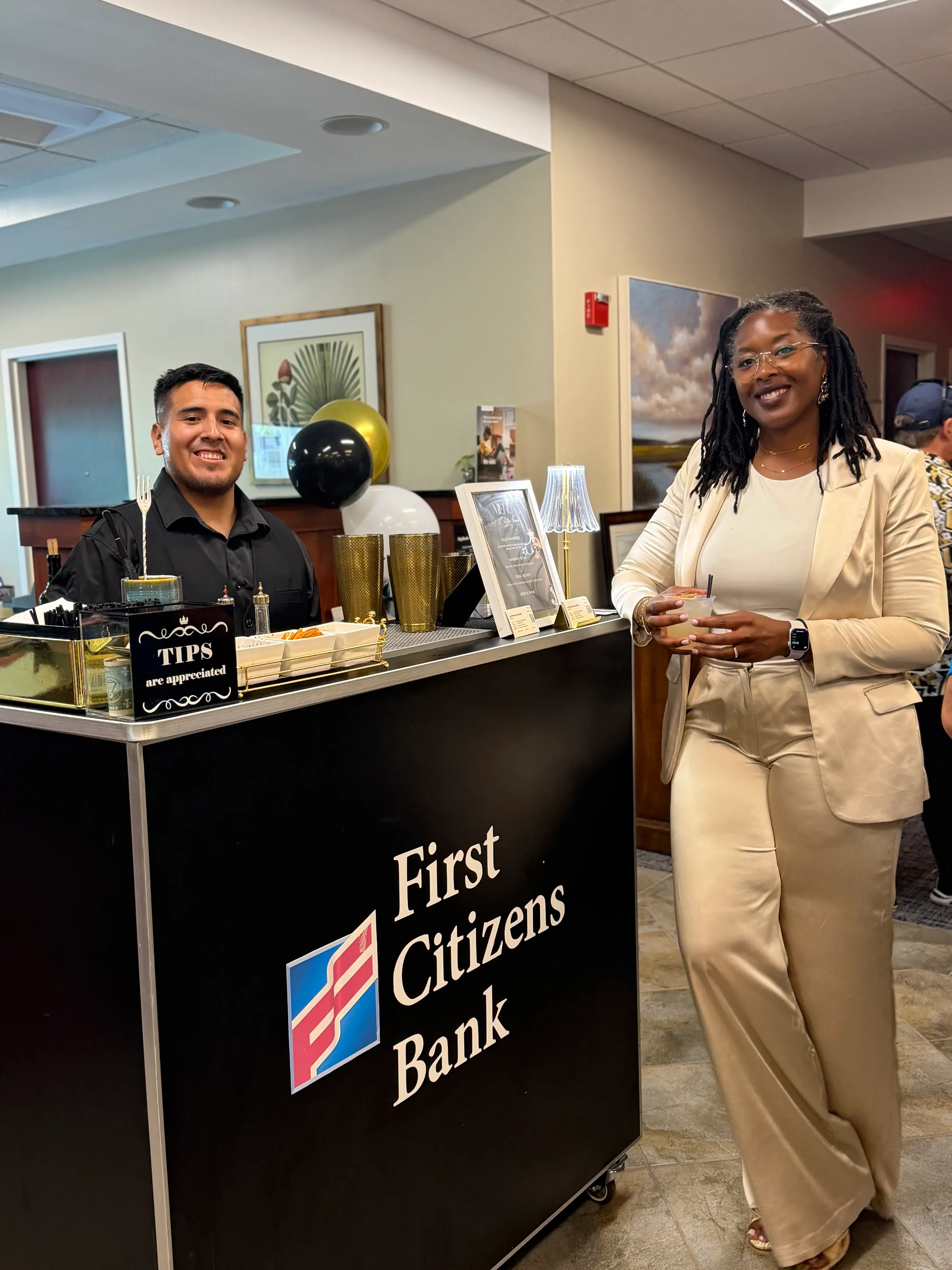 A woman in beige business attire standing next to a cash register at a bank, smiling and holding a cup, with a bank employee behind the counter.
