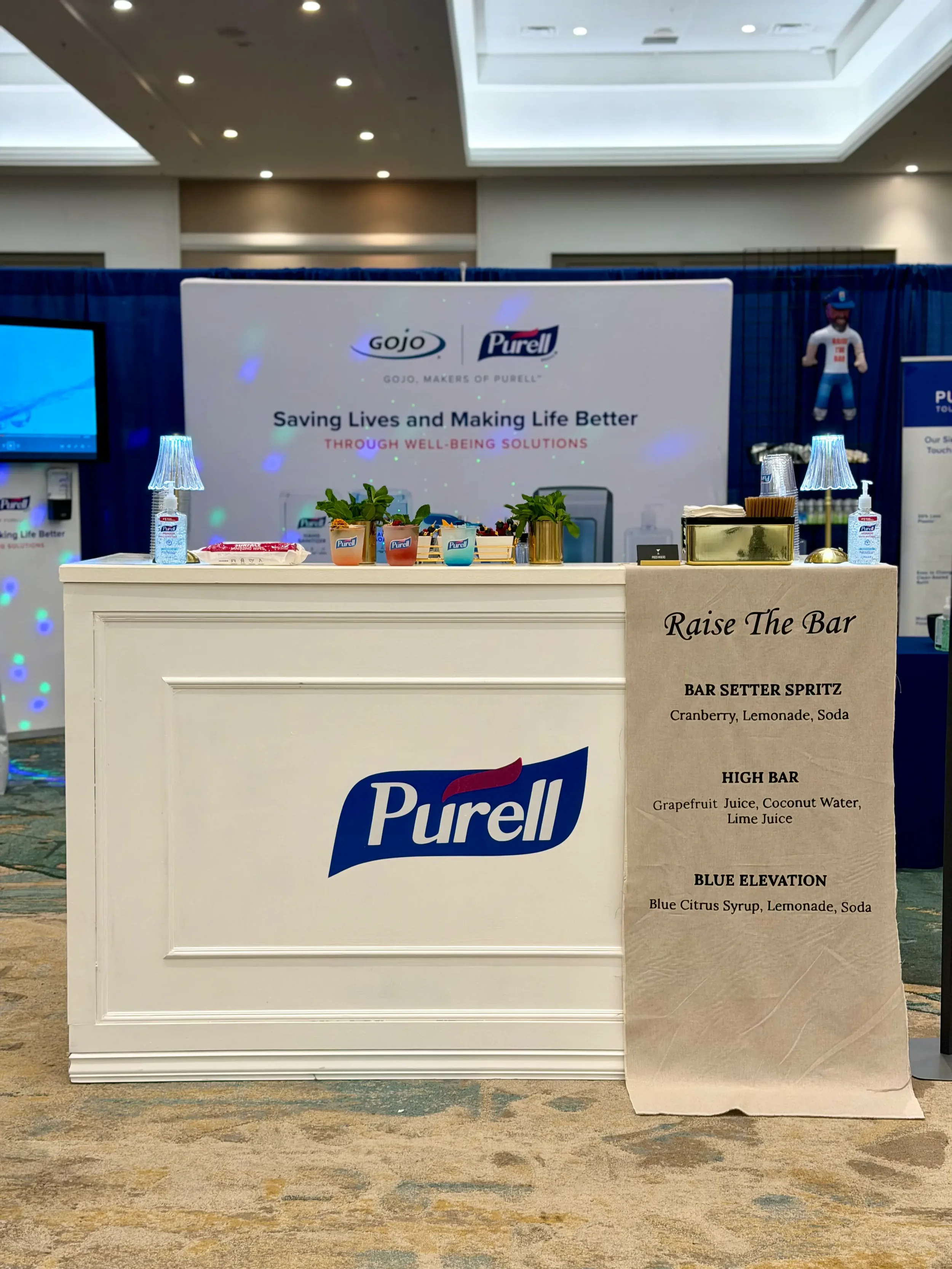 A booth with the Purell logo, promotional materials, and a sign listing different types of bar spritz drinks with ingredients. The booth has a white counter with a bottle of hand sanitizer, small potted plants, and some decorations. There is a large 