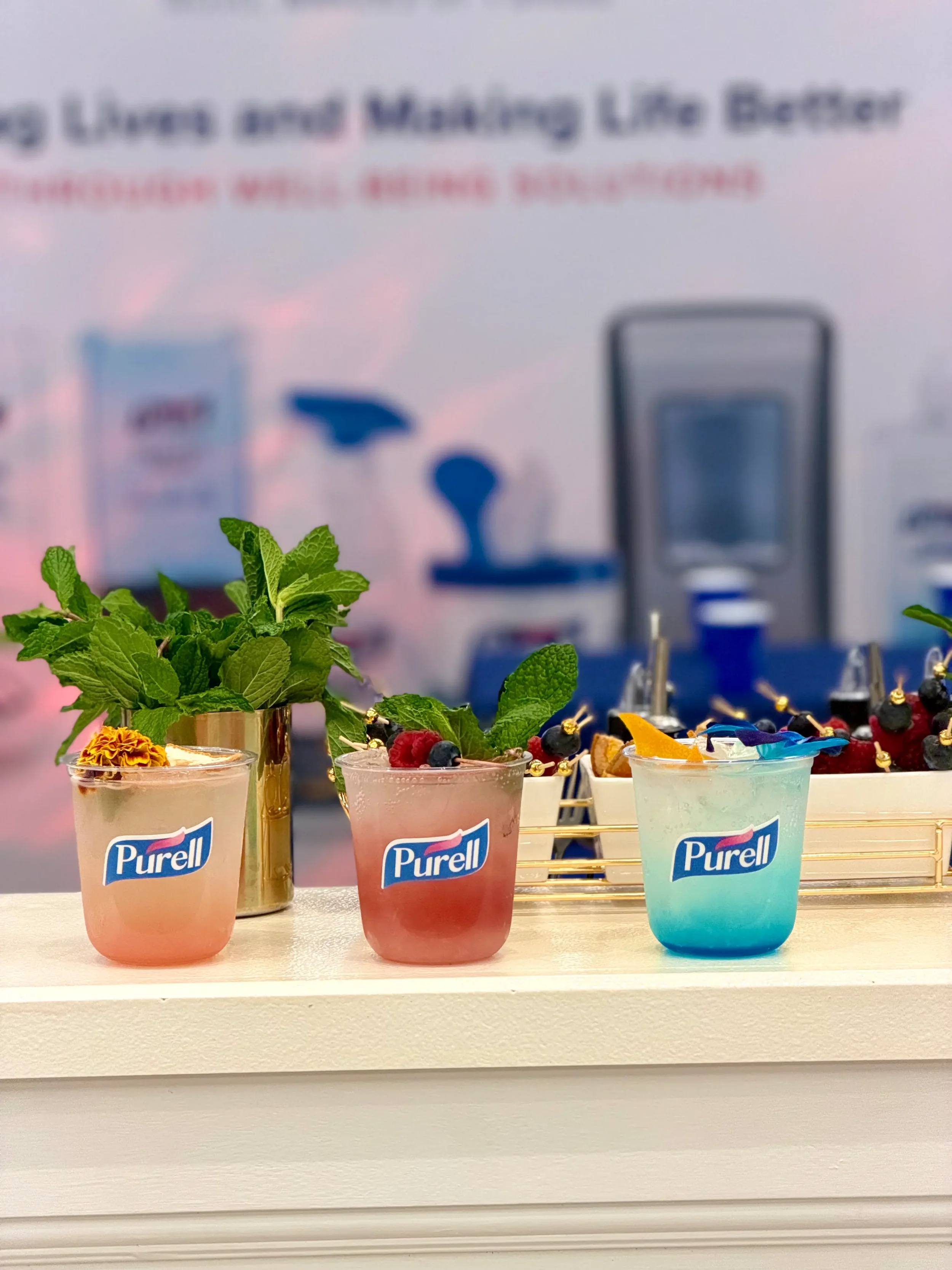 Three colorful drinks with mint, berries, and fruit garnishes, set on a white surface, with a blurred background featuring a sign and refill station.