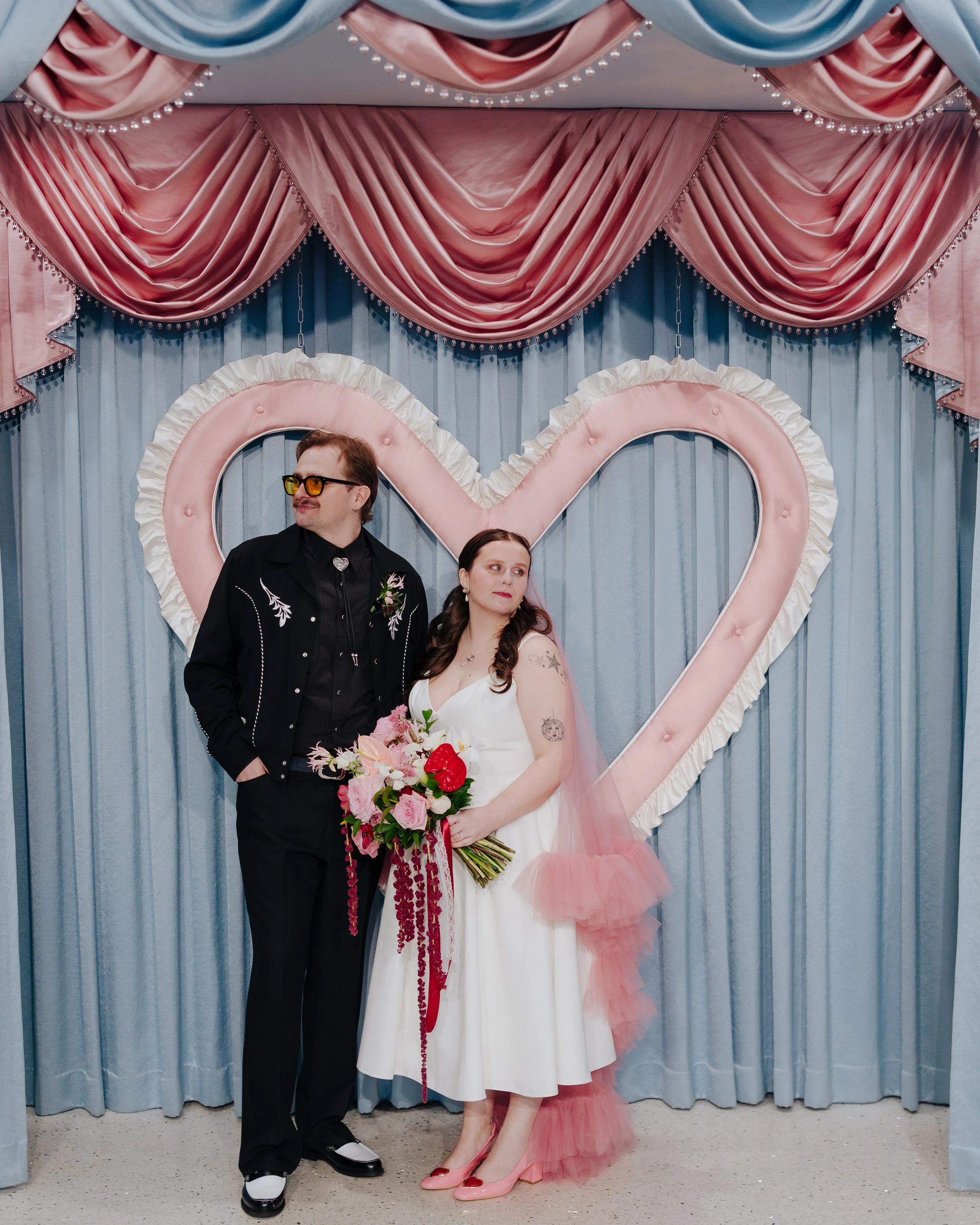 A couple standing in front of a large pink heart-shaped decoration with pink and blue drapes and pearl accents at a wedding or special event. The woman is holding a bouquet of flowers and wearing a white dress with pink shoes. The man is dressed in a