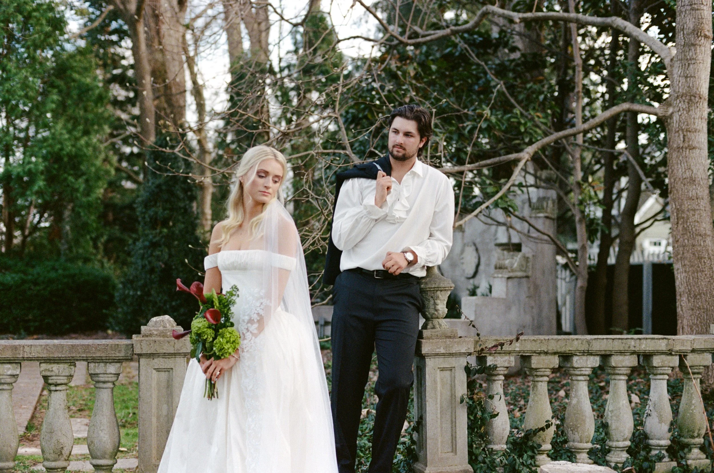 A bride in a white wedding dress holding a bouquet of flowers stands with a groom in a white shirt and dark pants at a garden with trees and ruins in the background.
