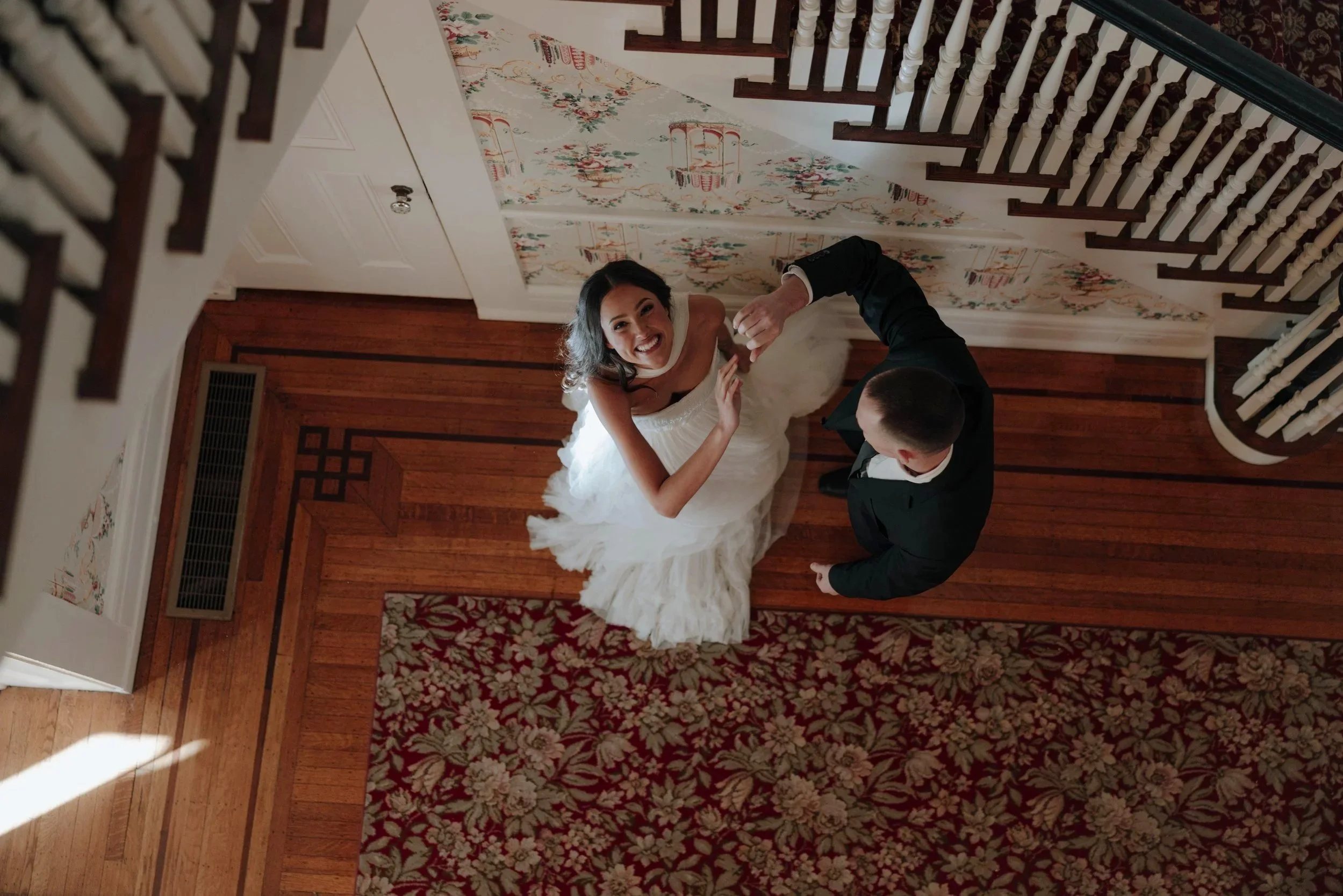 A bride and groom dancing on a wooden floor in a house, looking up at the camera with the bride smiling. DOCUMENTARY STYLE, EDITORIAL STYLE