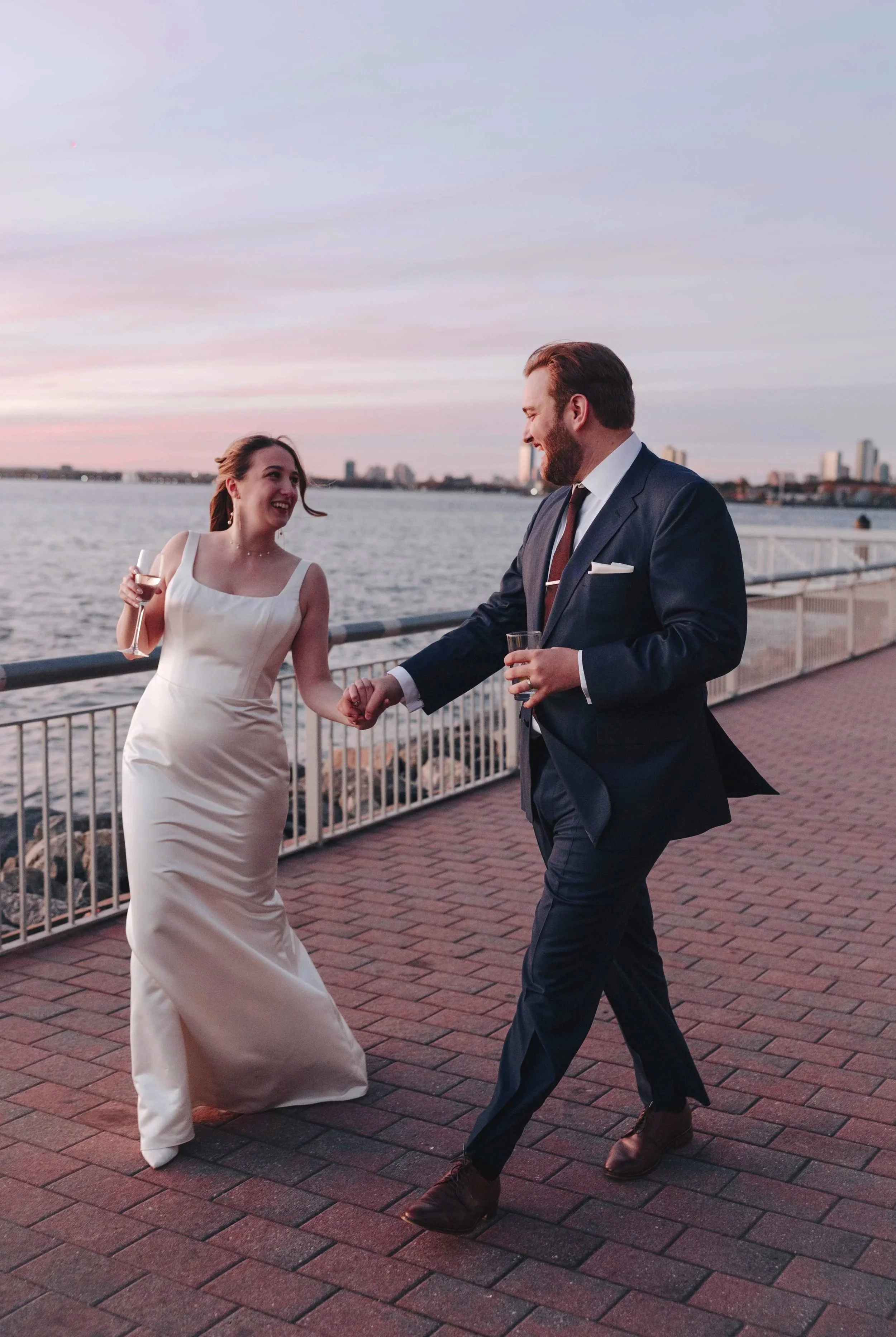 A couple in formal attire dancing and smiling by a riverside during sunset, with NYC city buildings in the background. DOCUMENTARY STYLE, EDITORIAL STYLE