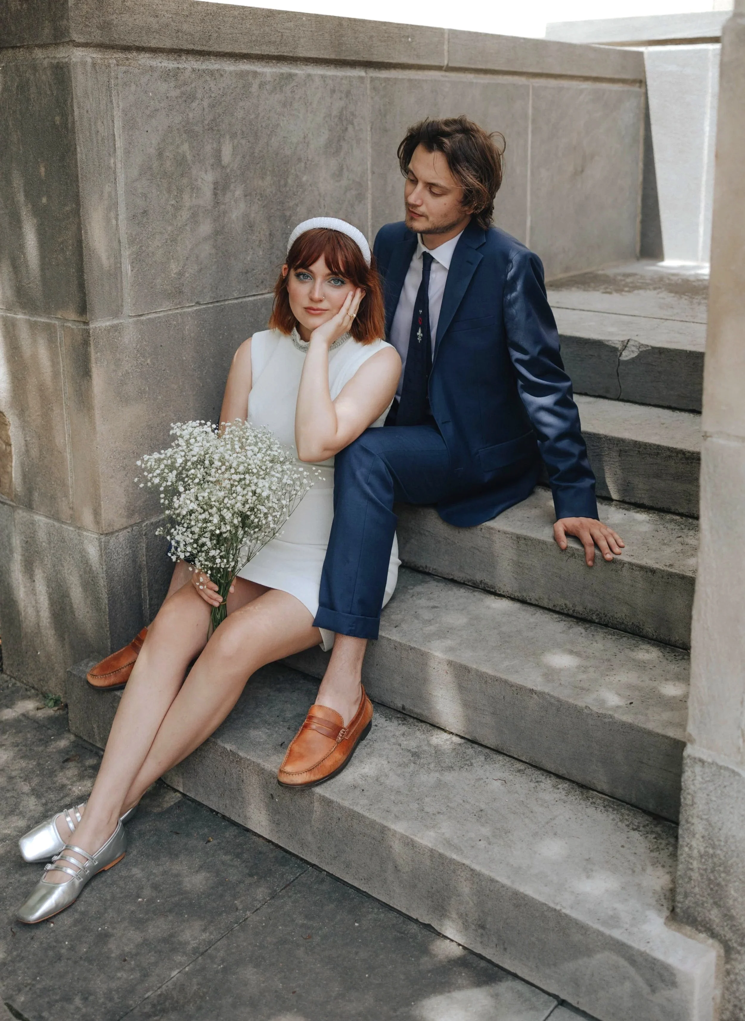 A woman in a white dress sitting on stairs holding a bouquet of flowers next to a man in a dark blue suit, both sitting on stone stairs. DOCUMENTARY STYLE, EDITORIAL STYLE