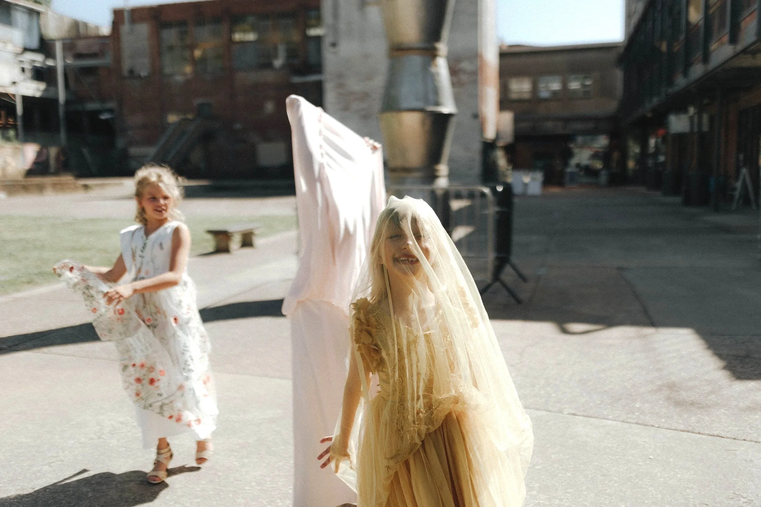 Two young girls in fancy dresses outdoors in a courtyard, with one girl smiling and wearing a veil, and another girl holding a floral dress, surrounded by urban buildings and a pipe as part of the background. DOCUMENTARY STYLE, EDITORIAL STYLE