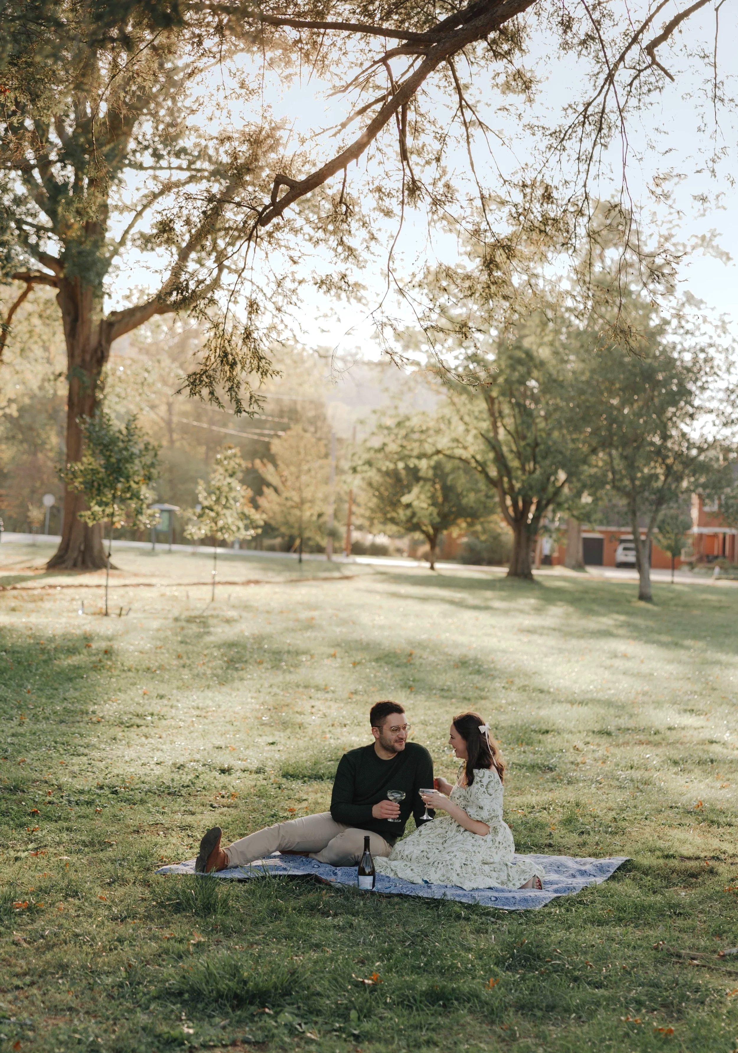 A couple sitting on a blanket having a picnic in a park with trees and sunlight in the background. DOCUMENTARY STYLE, EDITORIAL STYLE