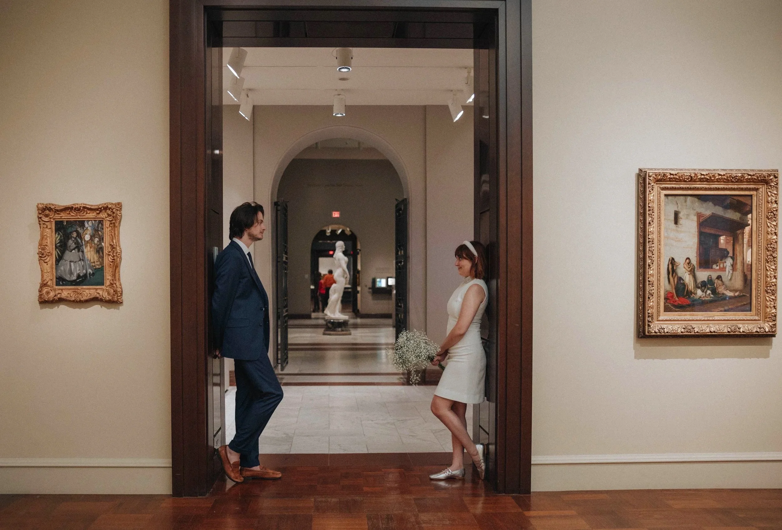 A man and a woman in a museum or art gallery talking to each other. The woman holds a bouquet of flowers and is dressed in a white dress, while the man is dressed in a blue suit. There are paintings on the walls and sculptures in the background. DOCU
