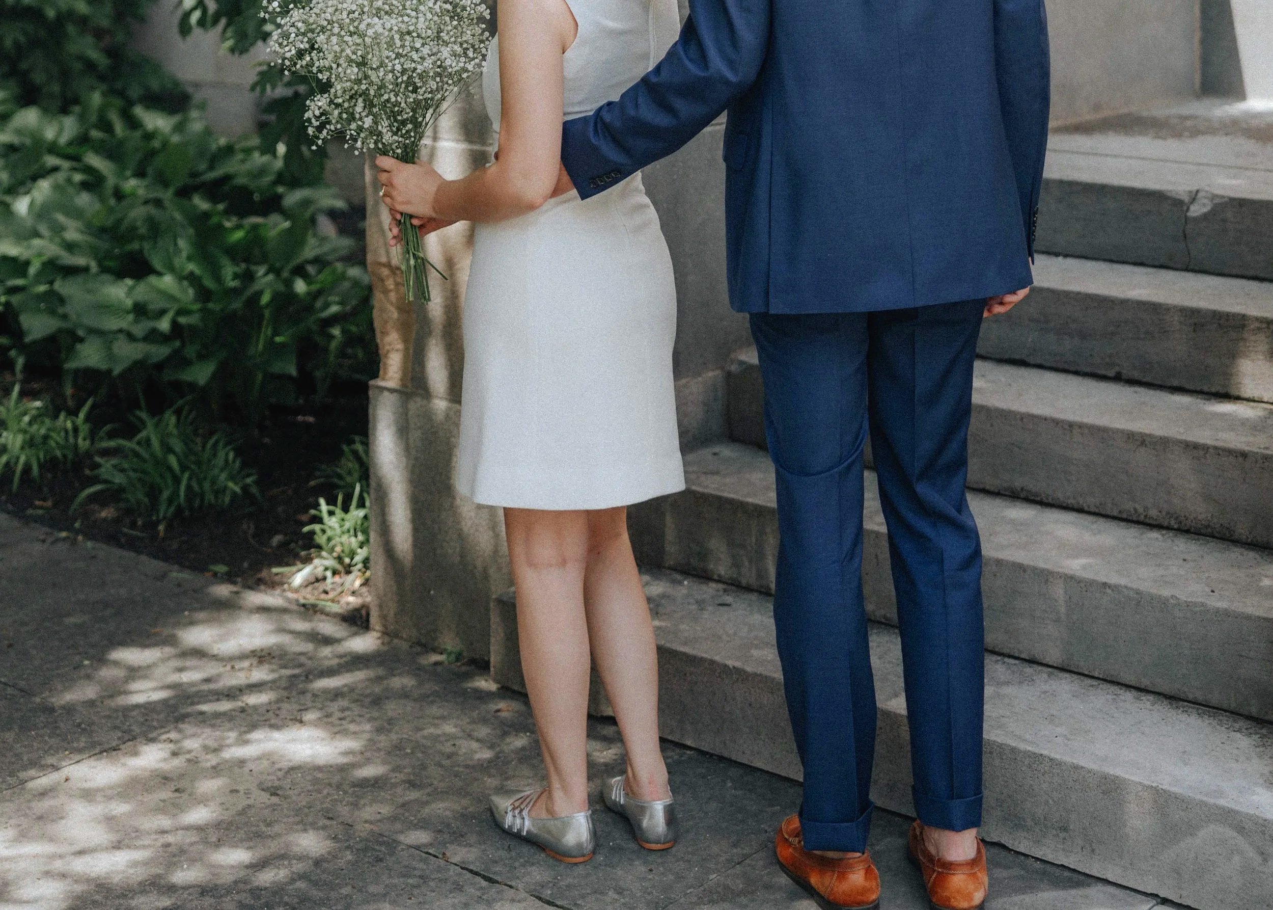 Couple dressed in wedding attire standing on outdoor steps, with woman holding a bouquet of white flowers.