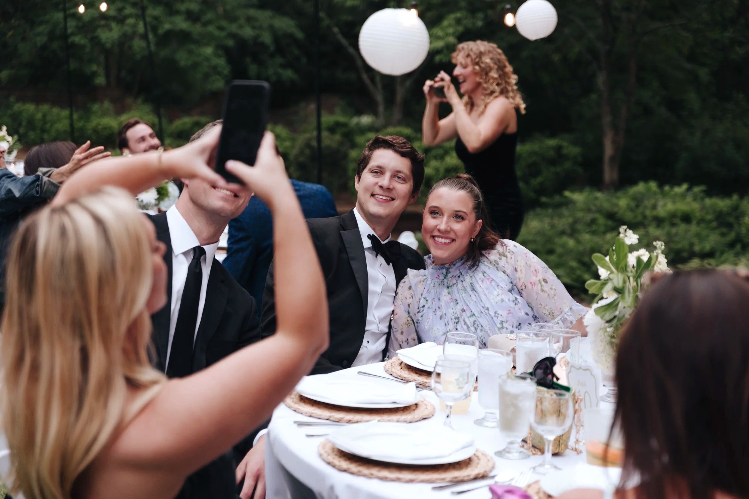 A group of people at an outdoor celebration, with a couple dressed in formal attire sitting at a table, smiling for a photo being taken by others, with hanging paper lanterns and lush greenery in the background.