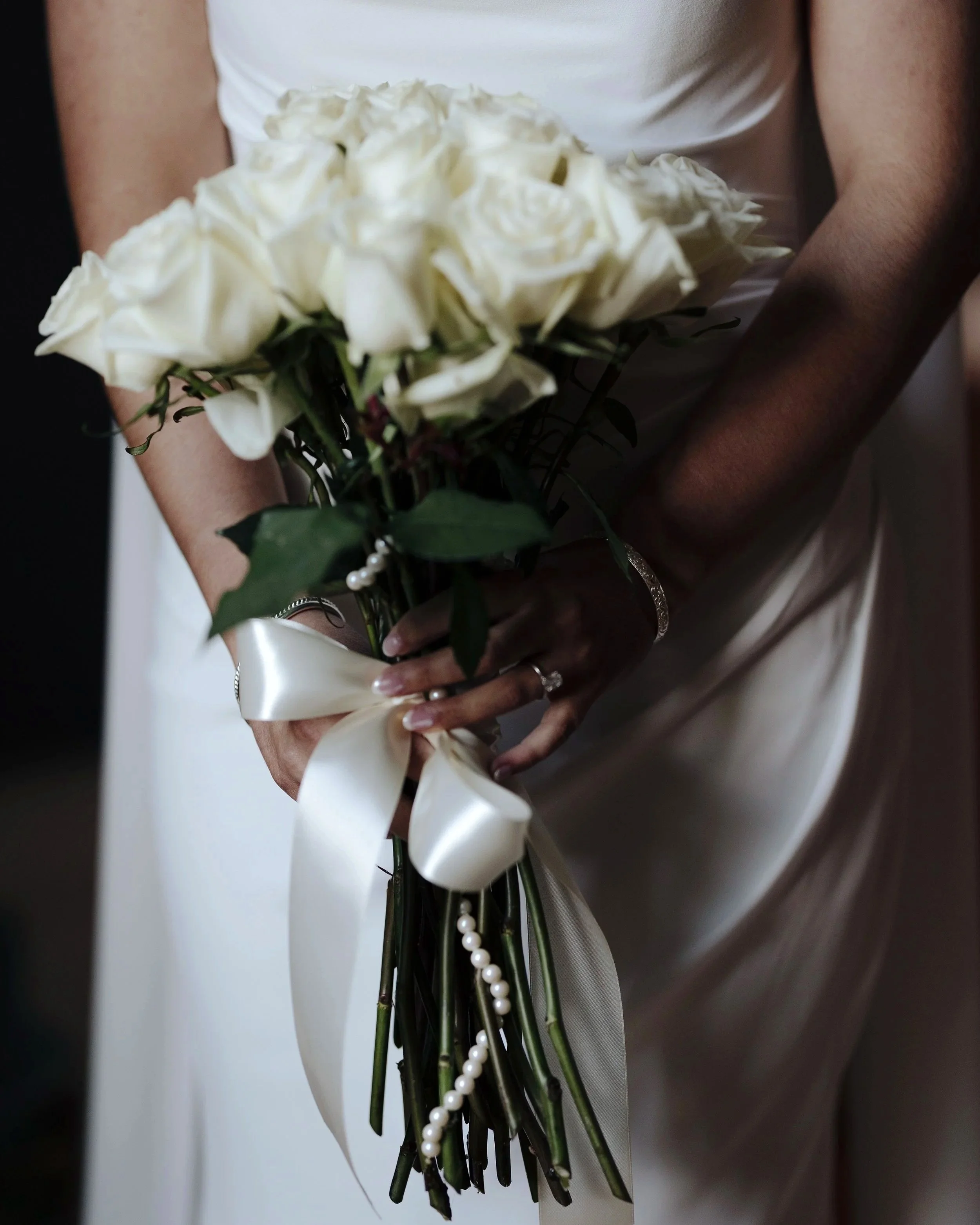 Person holding a bouquet of white roses decorated with a satin ribbon and pearl strands, dressed in a white gown.