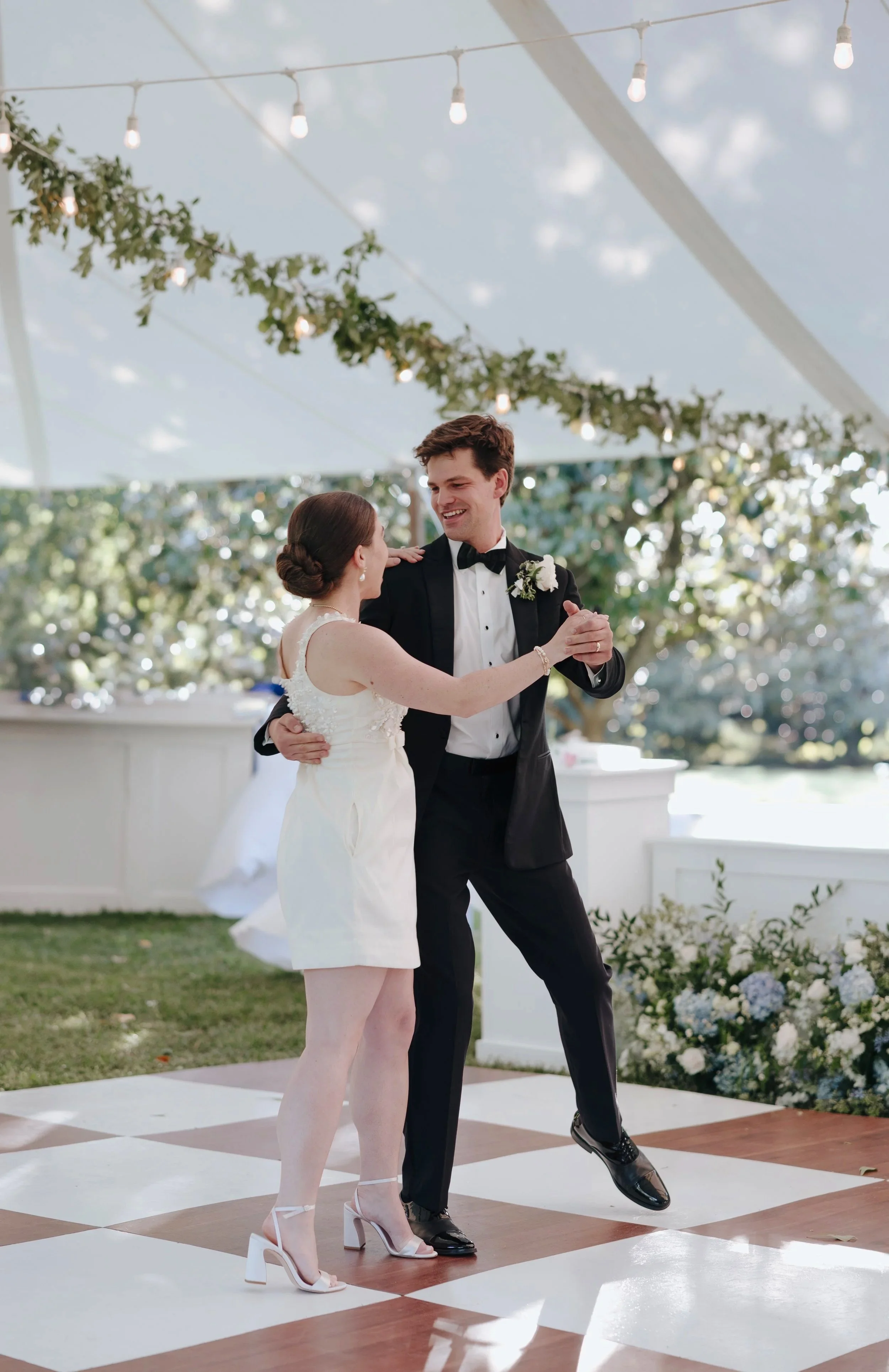 A bride and groom dancing at an outdoor wedding reception under a tent decorated with string lights and greenery, with a checkered dance floor and floral arrangements in the background. DOCUMENTARY STYLE, EDITORIAL STYLE