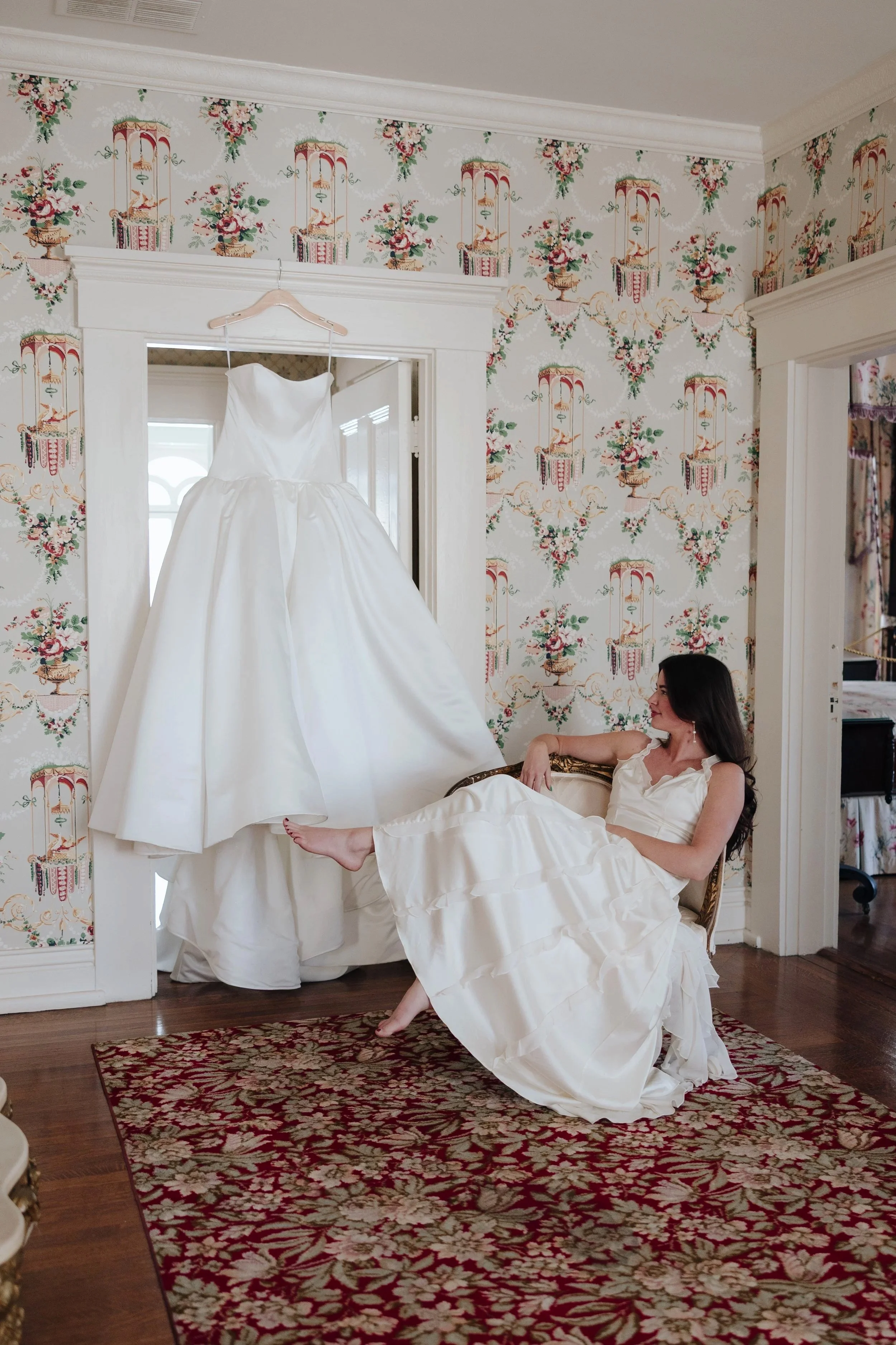 A woman in a white dress sitting on a vintage sofa, smiling and looking at a wedding dress hanging on a hanger in a room with floral wallpaper and a patterned rug.