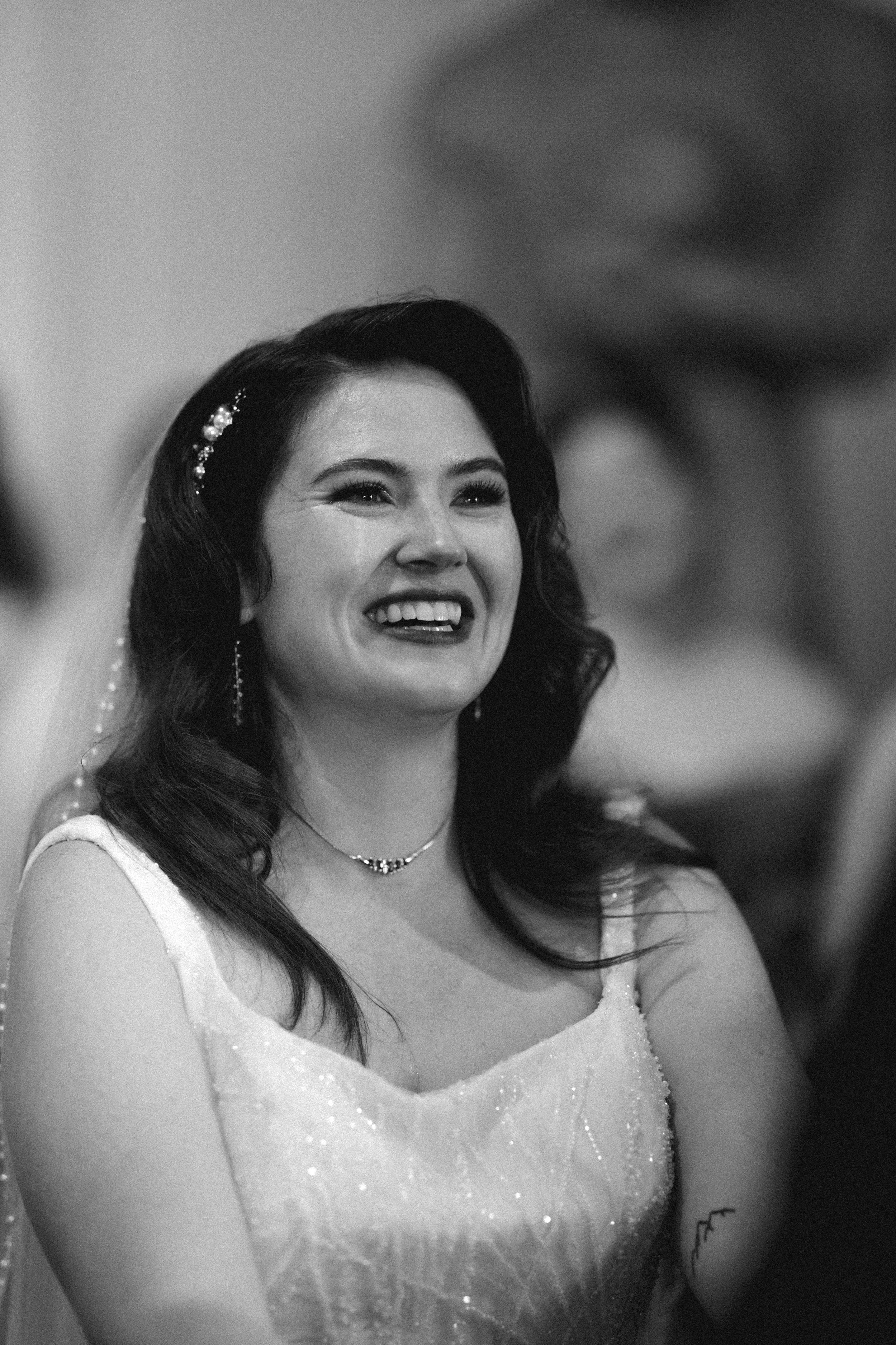 A woman smiling during a special event, possibly a wedding, wearing a dress and jewelry, with dark, wavy hair and a small tattoo on her arm. DOCUMENTARY STYLE, EDITORIAL STYLE