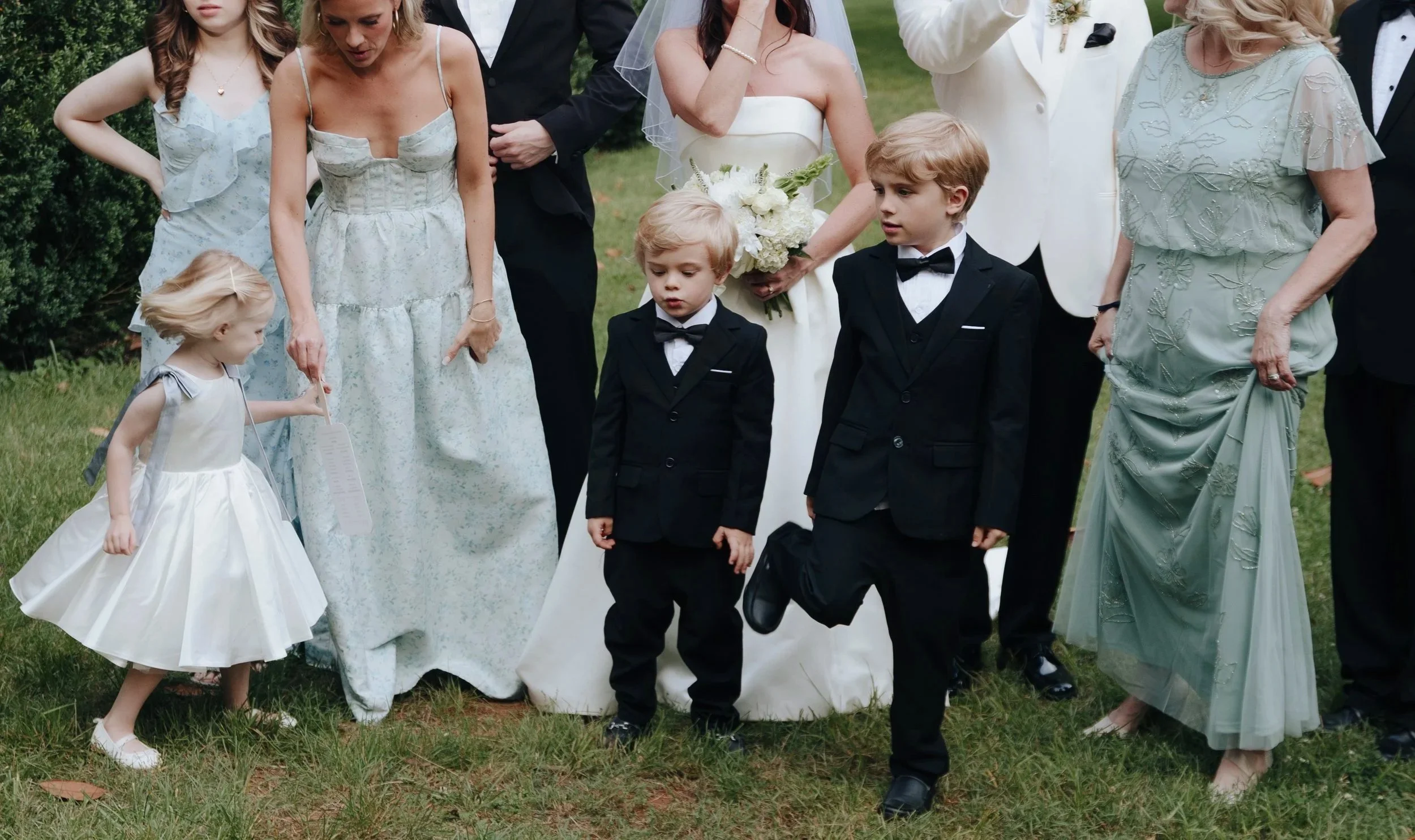 Group of people at a wedding, including children in formal attire, standing on grass outdoors, some looking down and others posing for the photo.