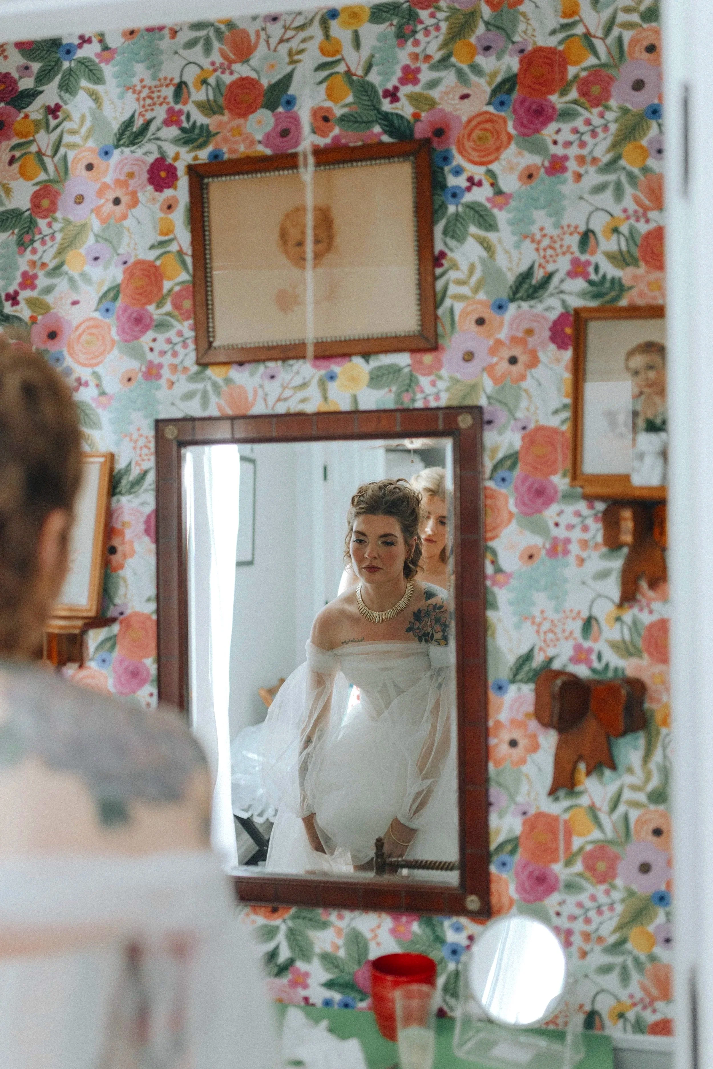 A woman in a white wedding dress is looking at herself in a mirror, with her reflection showing her adjusting her dress. The woman has dark hair styled up, and the room has floral wallpaper. DOCUMENTARY STYLE, EDITORIAL STYLE