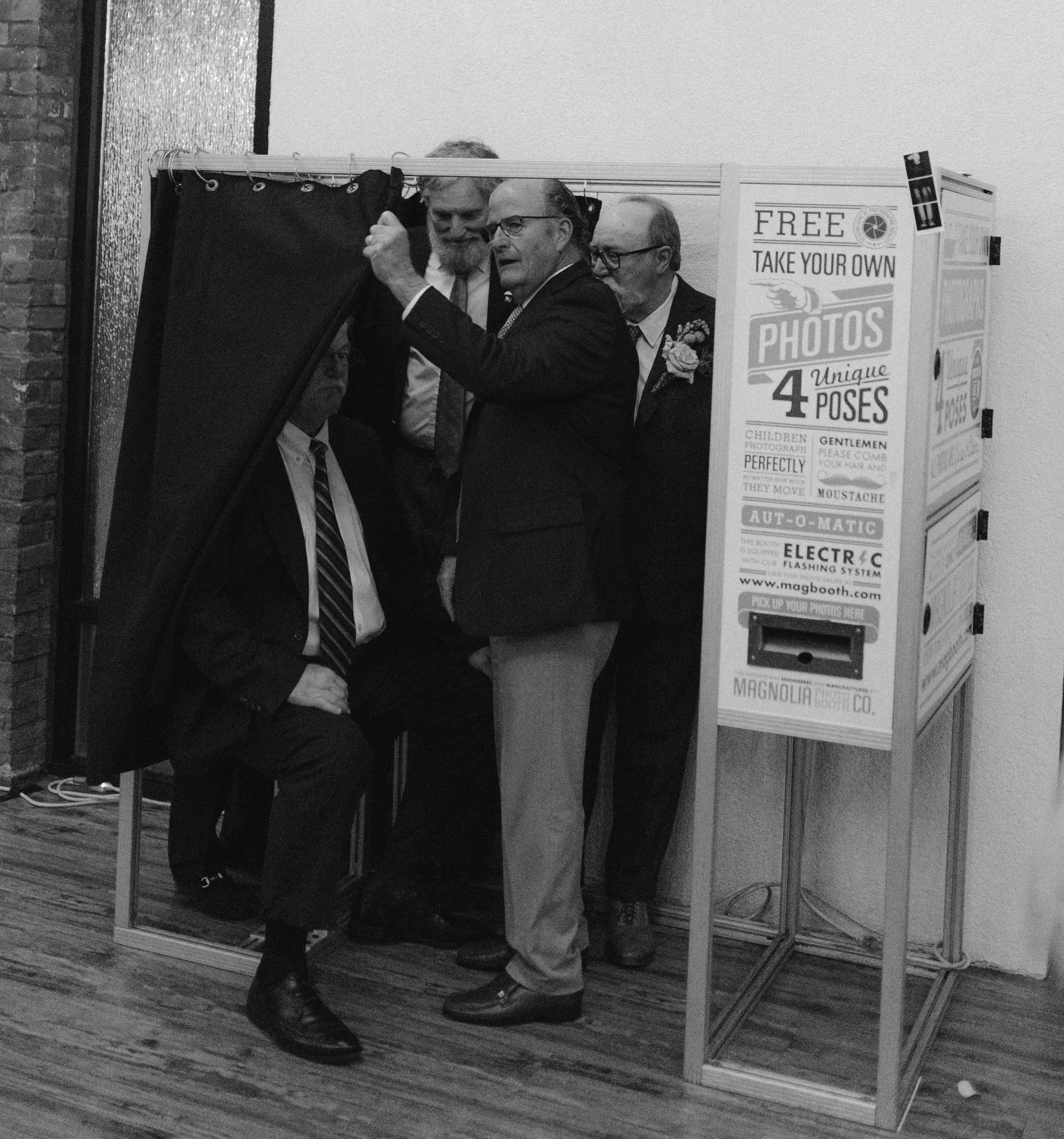 Four men standing around a photo booth with sign advertising free photo takes with four poses. One man is sitting in the booth while the others pose around him, with one adjusting or opening the booth curtain. DOCUMENTARY STYLE, EDITORIAL STYLE